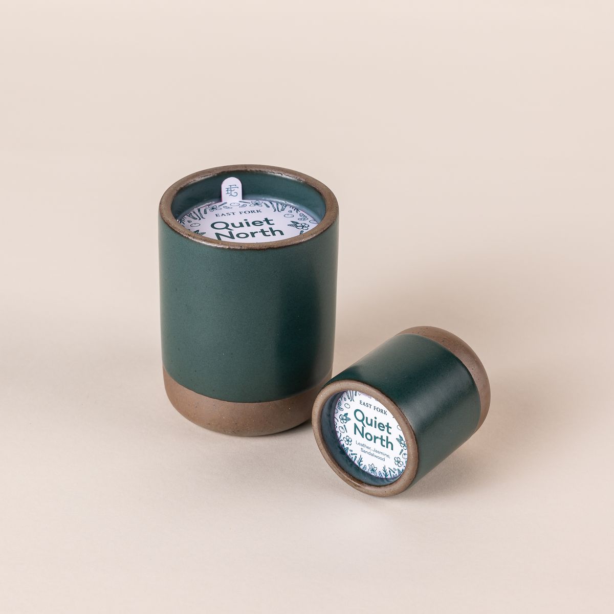 Large and small ceramic vessel in a deep dark teal color with candle inside. On top of each is a packaging label sitting on top that reads "Quiet North". The small candle lays on its side.