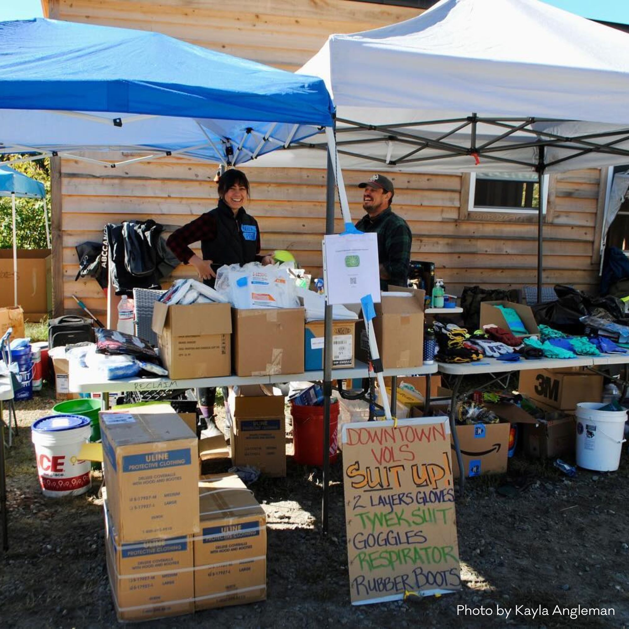 2 people are smiling and outside in pop up tents with lots of supplies and signage that reads "Downtown Vols" with rules to suit up for cleanup.