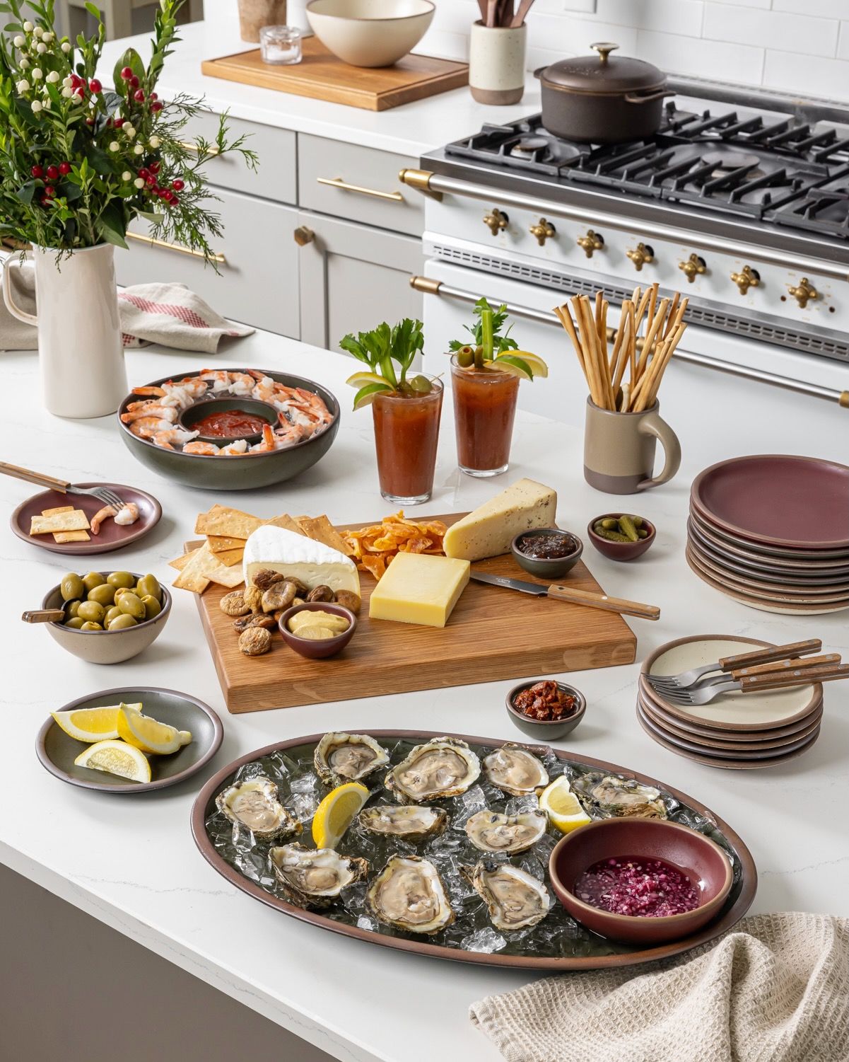 A kitchen island spread with a ceramic oval platter filled with oysters, a charcuterie board, bloody marys, shrimp platter, and plates.