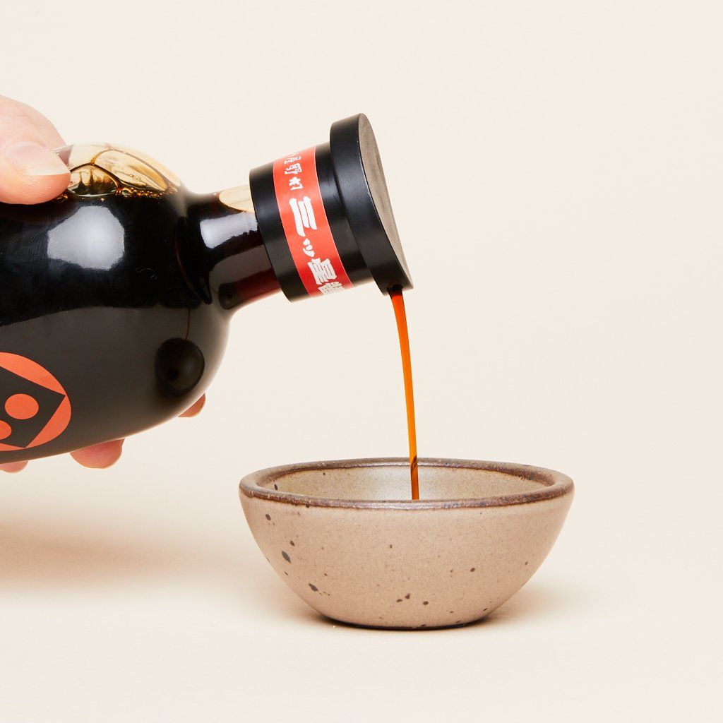 A stream of dark brown liquid pours from a bottle of soy sauce into a small brown bowl