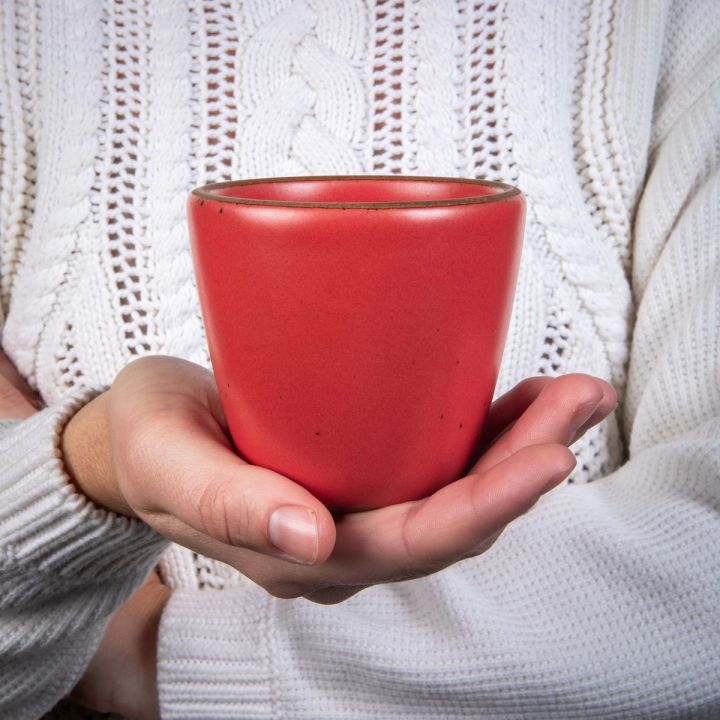 A hand holds a short cup that tapers out to get wider at the top in a bold red color featuring iron speckles.