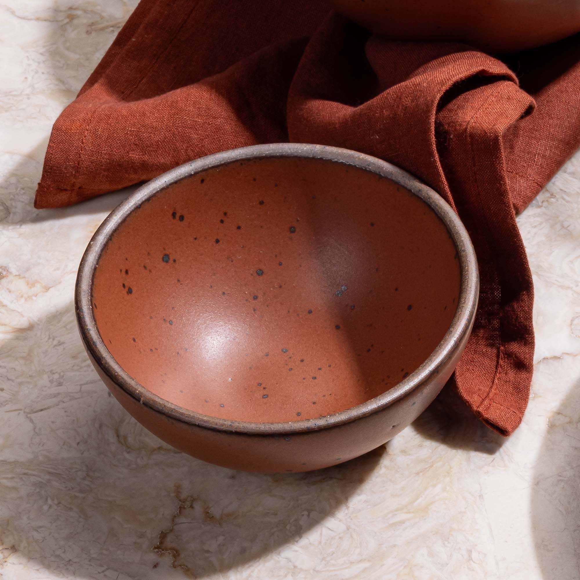 A small dessert sized rounded ceramic bowl in a cool burnt terracotta color with a napkin the same color.