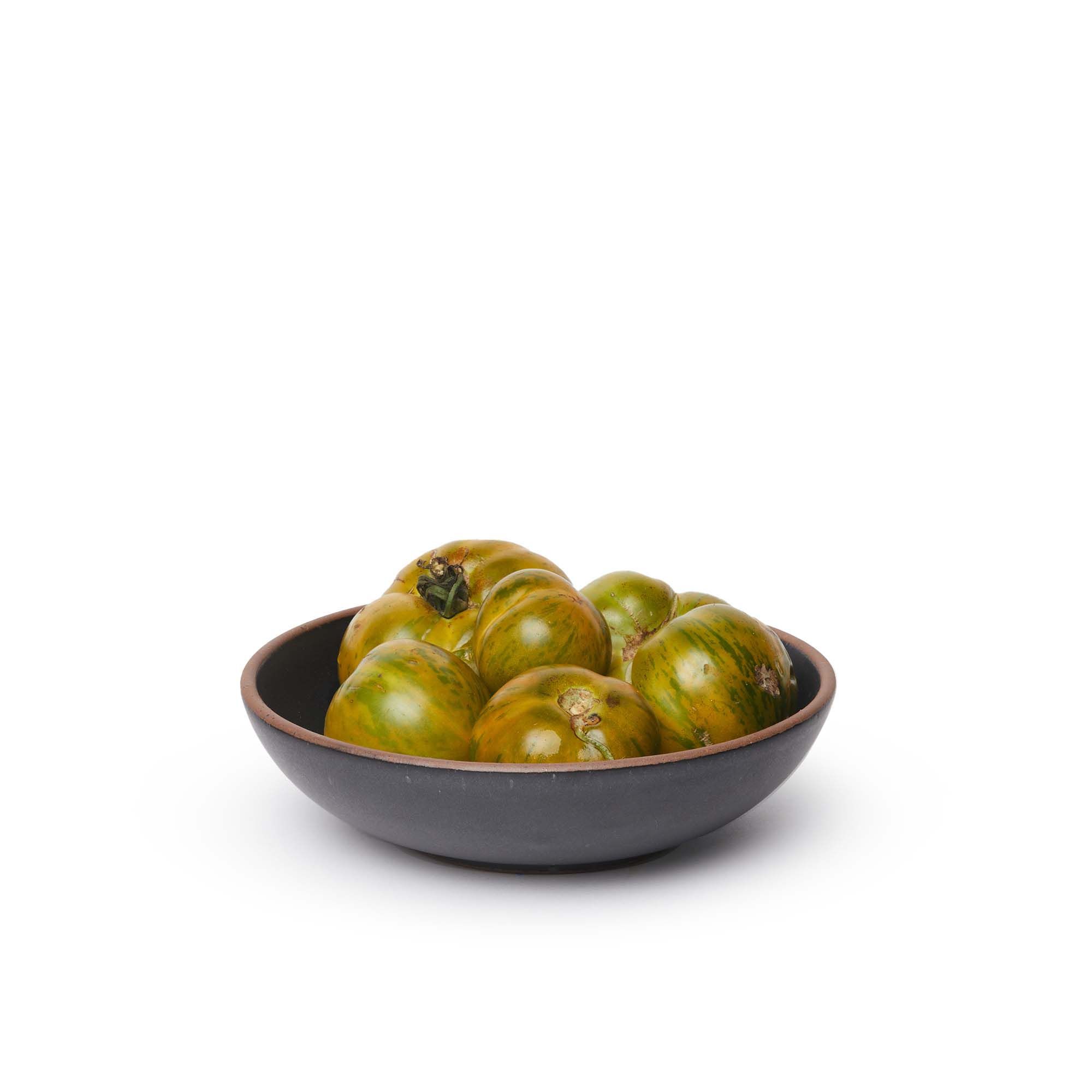 A dinner-sized shallow ceramic bowl in a graphite black color featuring iron speckles and an unglazed rim, filled with tomatoes