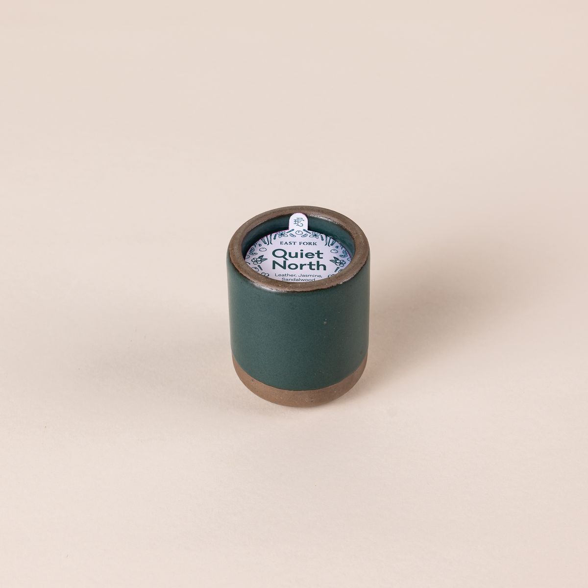 Small ceramic vessel in a deep dark teal color with candle inside. On top is a packaging label sitting on top that reads "Quiet North"
