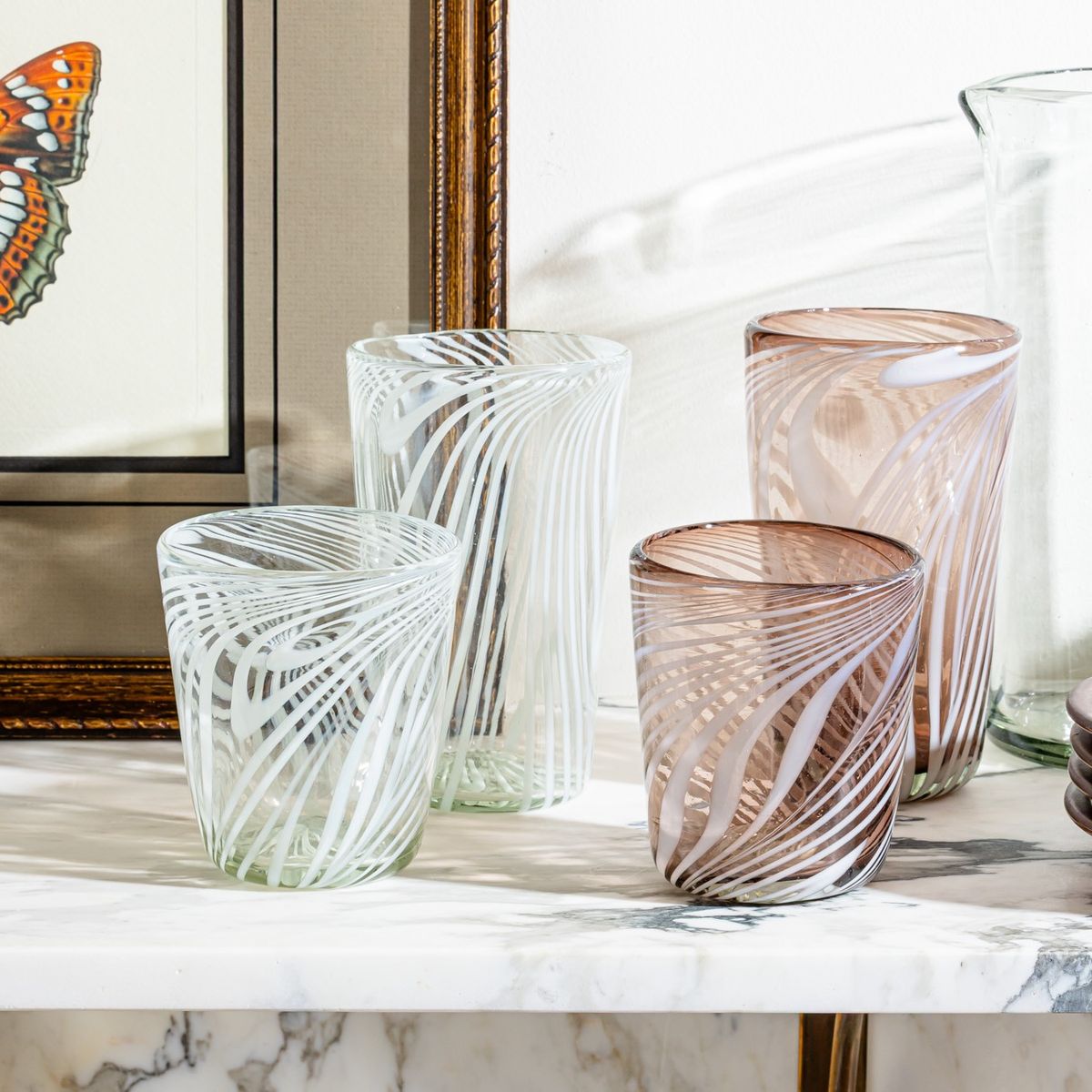 Swirled glass tumblers in clear and amber tones displayed on a marble surface with framed art in the background.