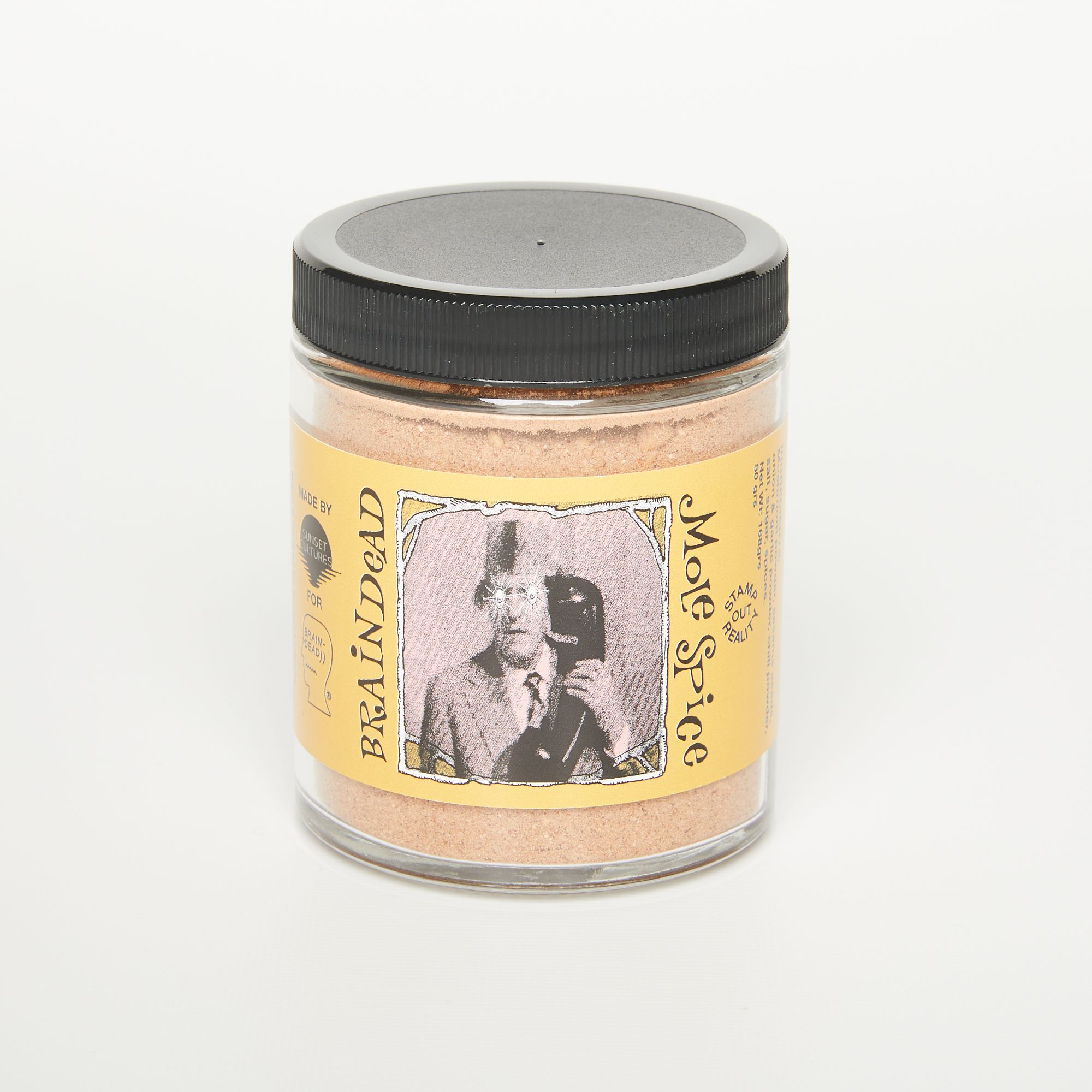 A short cylinder jar with a black screw cap and a butter yellow label that reads "Braindead Mole Spice" and has a photo of a man with a large phone.