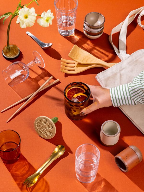 An artful flat lay of a hand reaching for a glass surrounded by tiny ceramic cups, more glassware, chopsticks, a cookie stamp, tote bag, and more