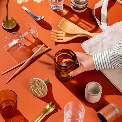 An artful flat lay of a hand reaching for a glass surrounded by tiny ceramic cups, more glassware, chopsticks, a cookie stamp, tote bag, and more