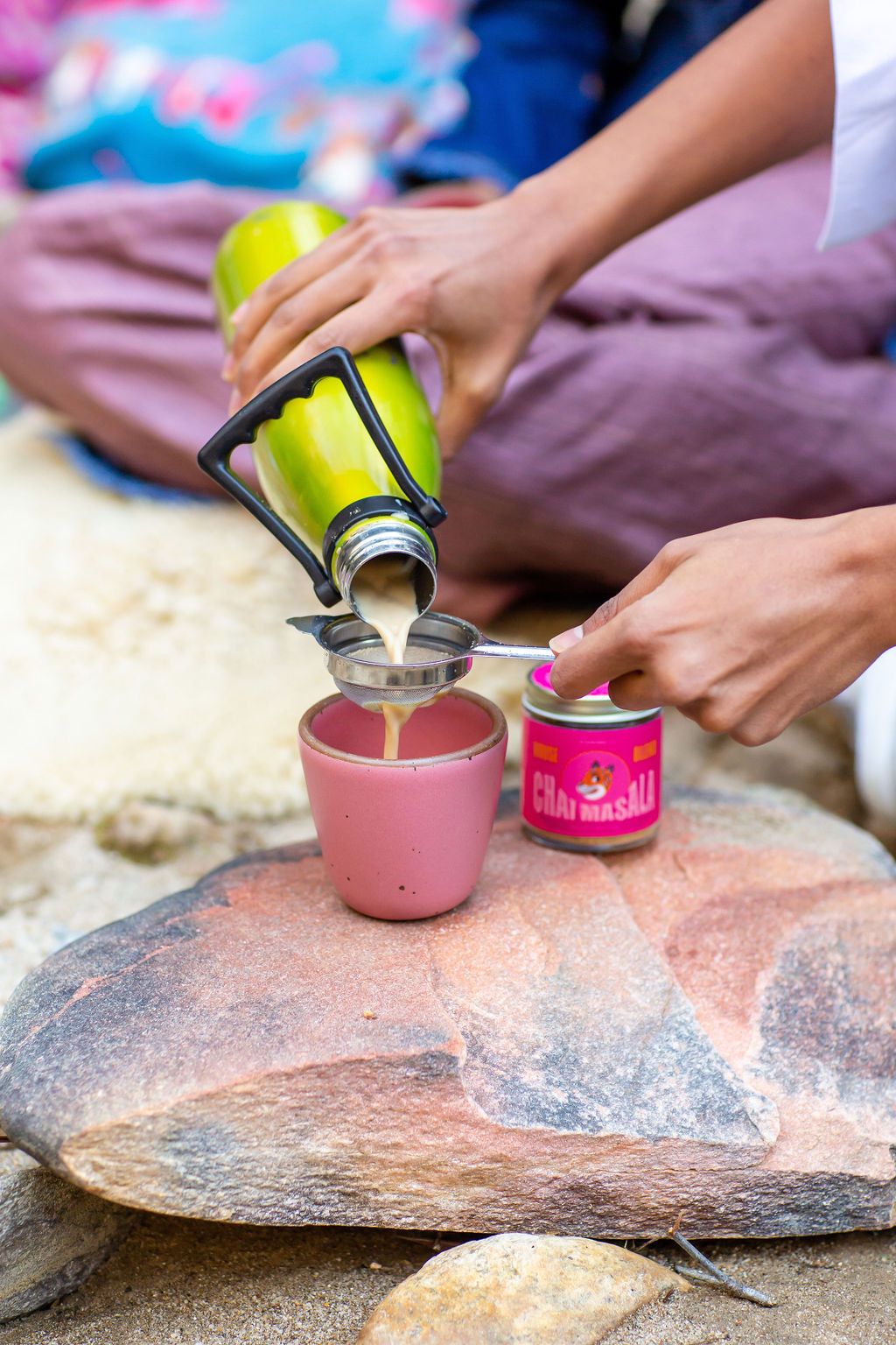 A tan and milky beverage pours from a bright green thermos into a hot pink cup on a salmon colored rock alongside the Hungry River