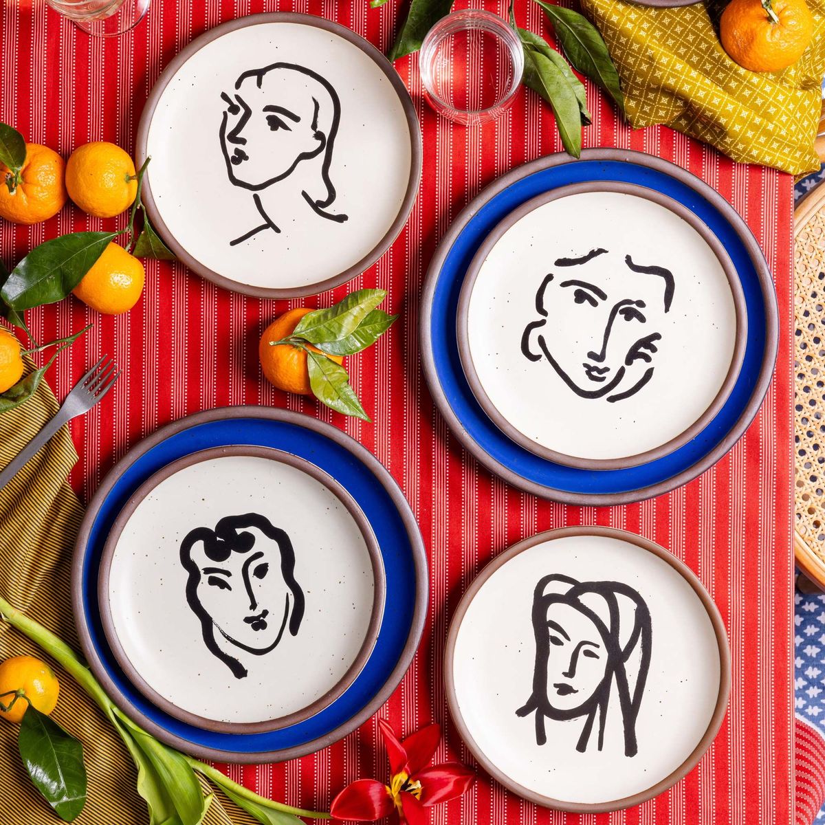 Four ceramic medium-sized plates in a warm off-white color with unglazed rim, featuring black aquatint portrait artworks by Henri Matisse. The plates are on a table with a red tablecloth, flowers, oranges, and more.