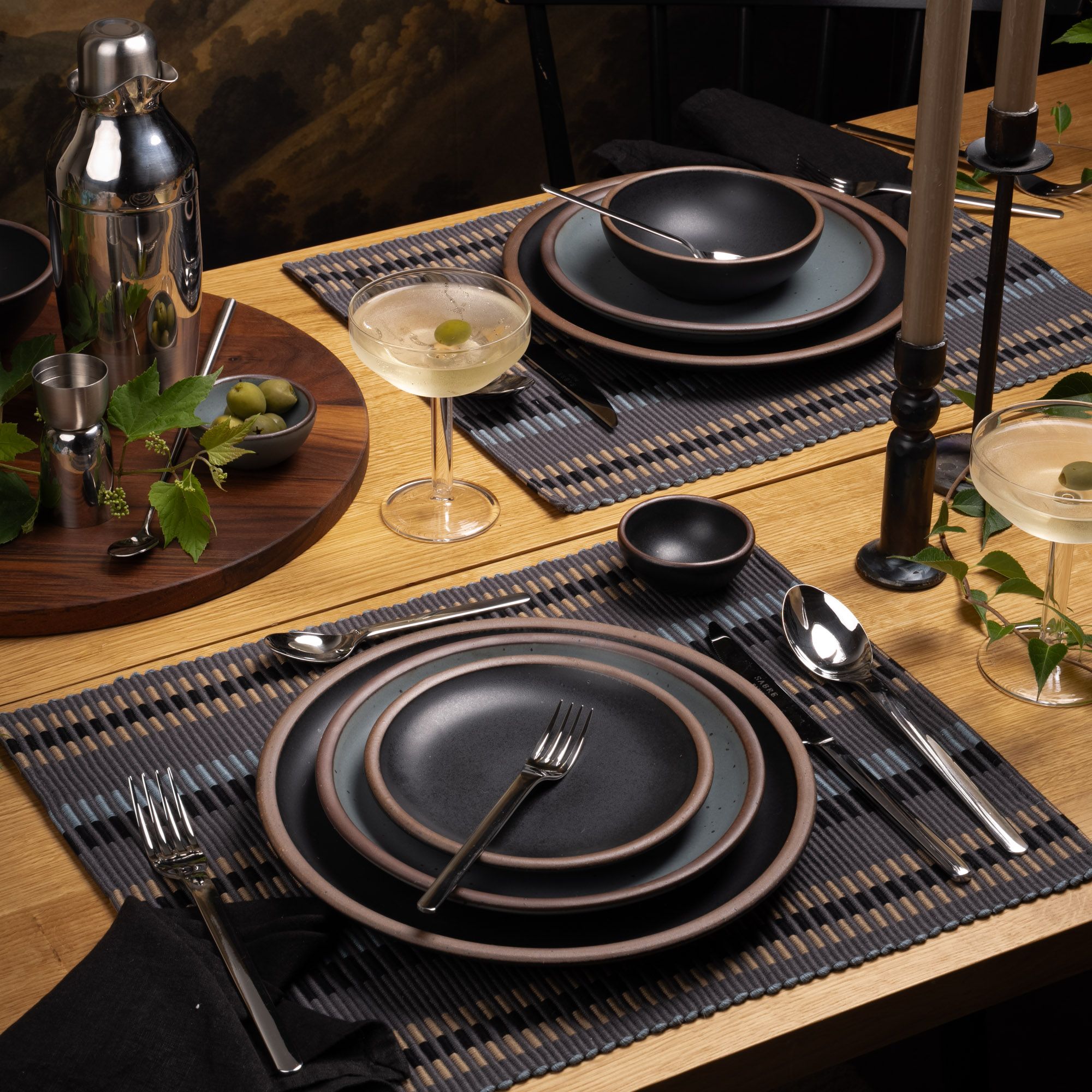 Elegant table setting with matte black stoneware, martini coupes, and a steel shaker on a wooden table with cool toned woven placemats.