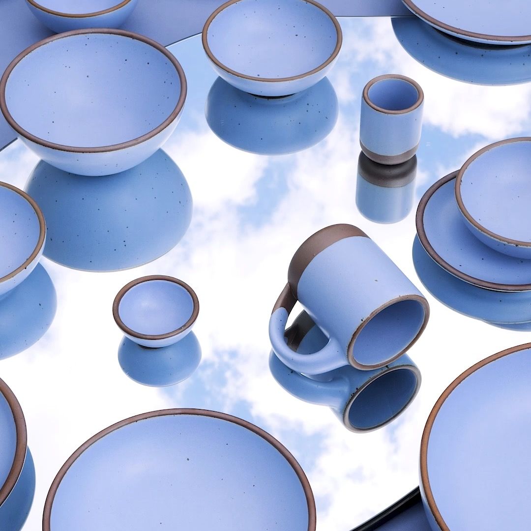 Ceramic plates, bowls, and a mug in a periwinkle color sit on a mirror that reflects a cloudy sky