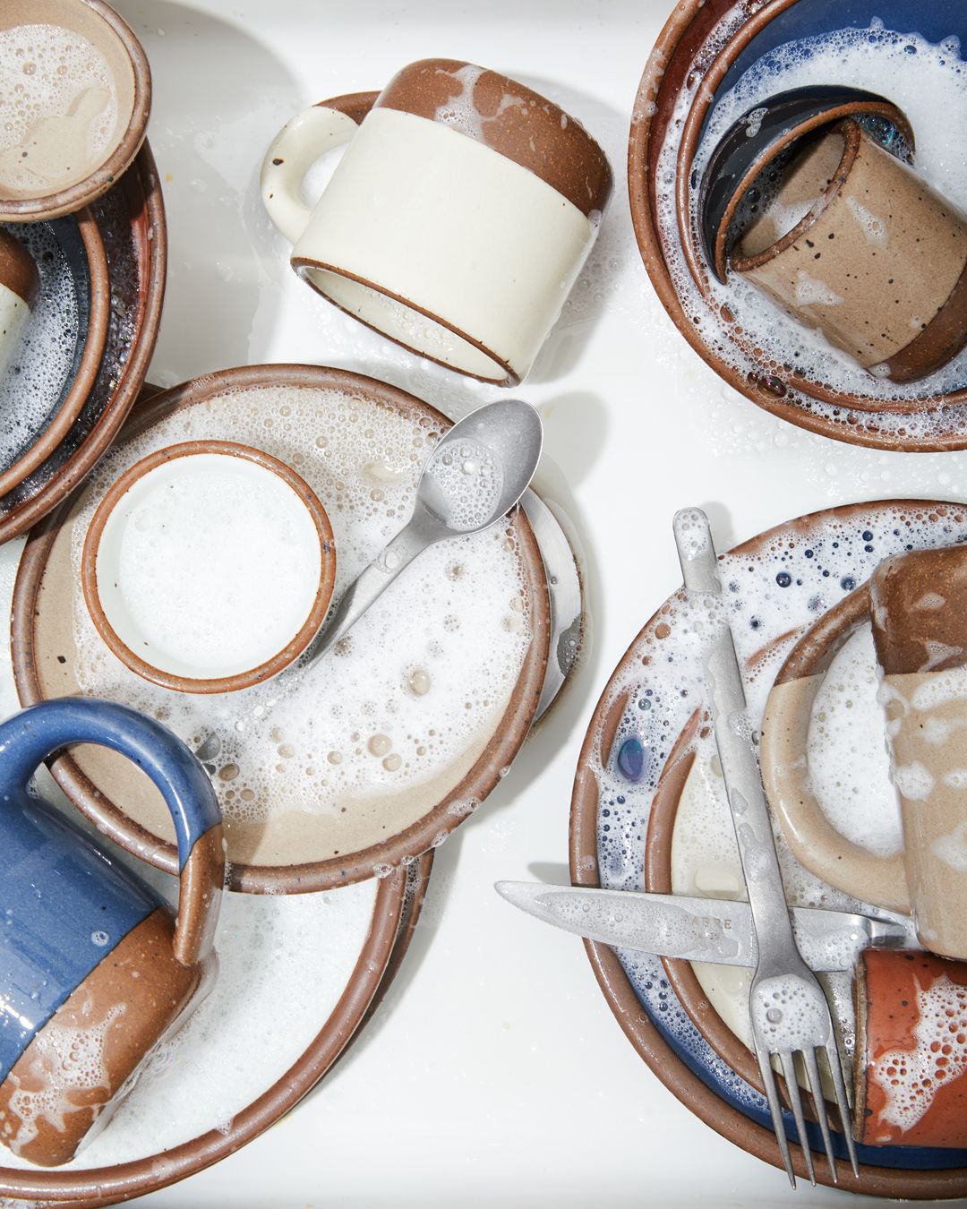 In a sink are various ceramic dishes, mugs, and silverware with sudsy water
