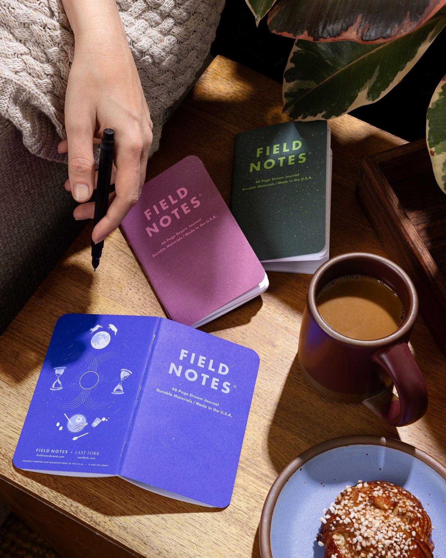 In a cozy setting, a hand is holding a pen and a mug in a deep plum color with 3 pocket sized journals next to it