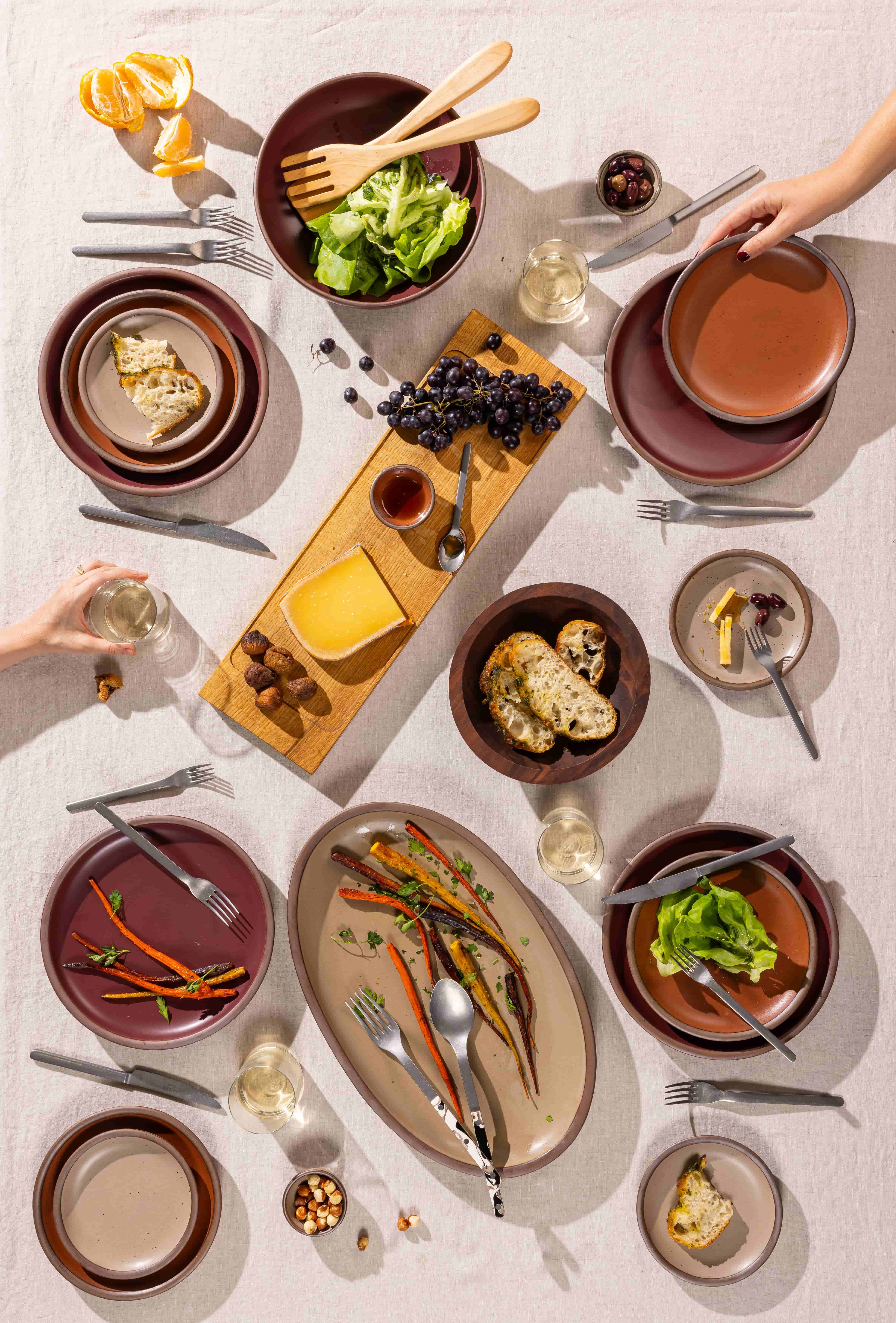 An overhead view of a table filled with several ceramic plates, an oval platter, wooden bowls, and a rectangular charcuterie board, along with silverware and wine glasses.