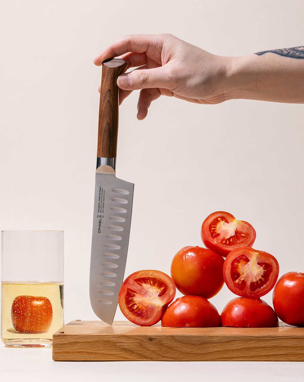 A hand holds up a santoku knife with a sharp steel blade with dimples and a beveled dark wood handle. The knife is being held upside down on a cutting board with a tower of tomatoes both full and sliced.