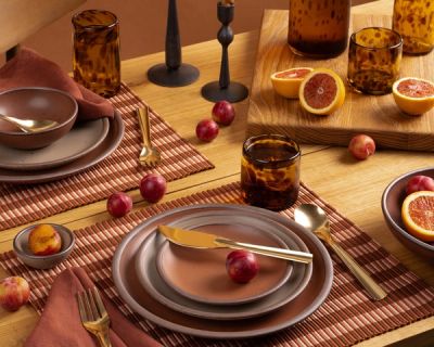 Rust-toned table setting with stoneware plates, amber glasses, plums, and blood oranges on striped placemats and a wood table.
