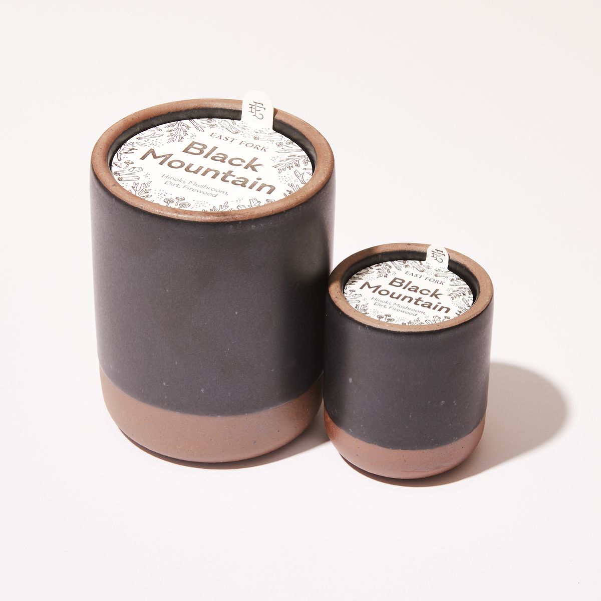 Small and large ceramic vessel next to each other in dark charcoal color with candles inside each. On top of each is a packaging label sitting that reads "Black Mountain"