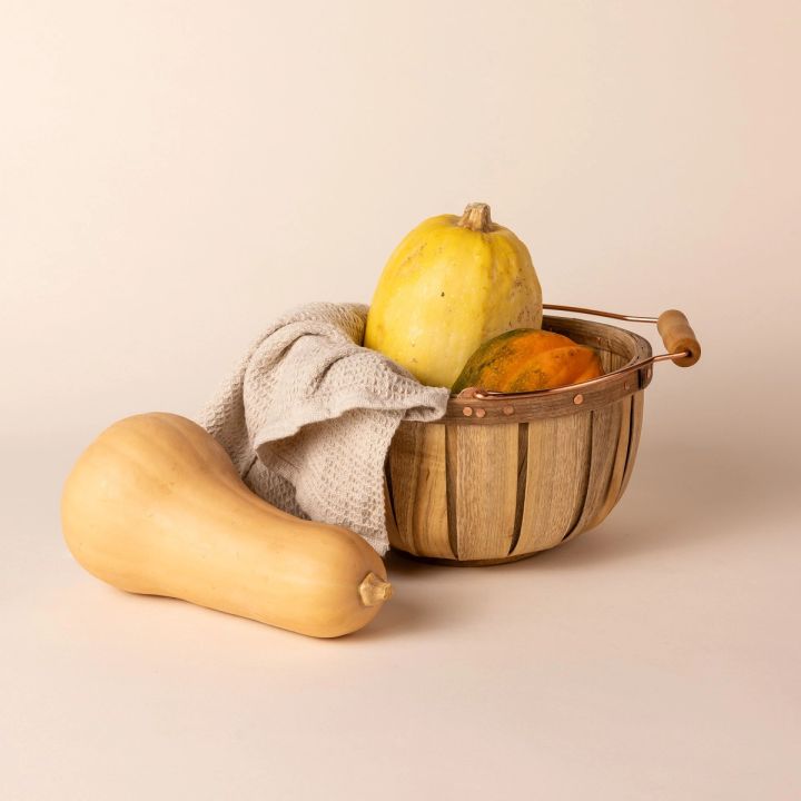 A short large tall round rustic wooden basket with a metal and wood handle is filled with yellow and orange squash along with a natural kitchen towel. A large squash leans on the side of the basket.