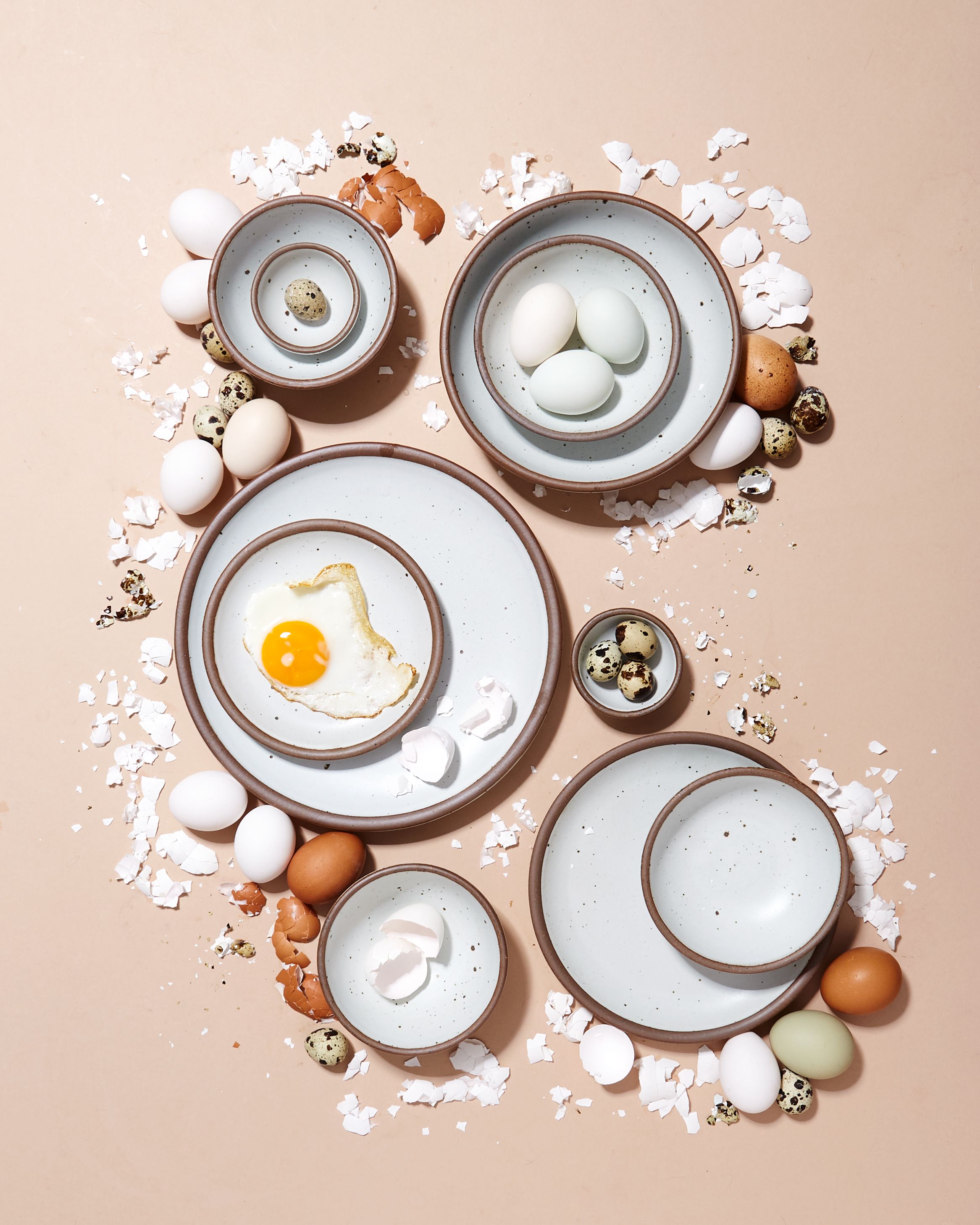 An overhead view of ceramic bowls and plates in various sizes in soft cool white and surrounded by eggshells and different kinds of eggs.