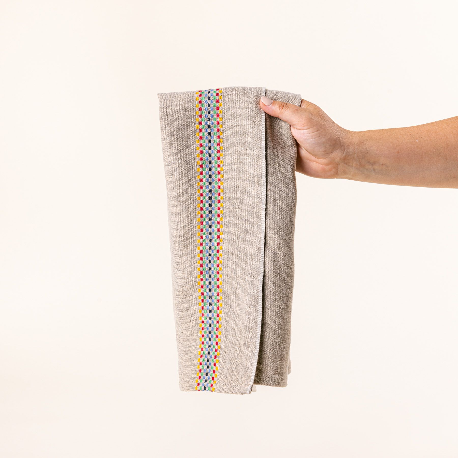 A hand holds a folded linen towel in a beige color with a multicolor checkerboard stripe down the side