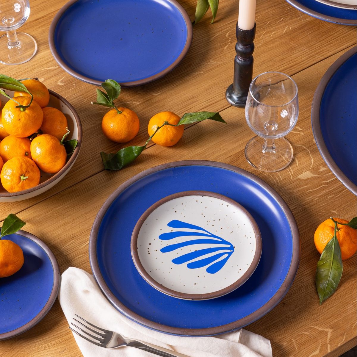On a table are place settings including large plates in a bold cobalt color with a small dessert-sized plate with a frond-like cut-out, with oranges and a wine glass.