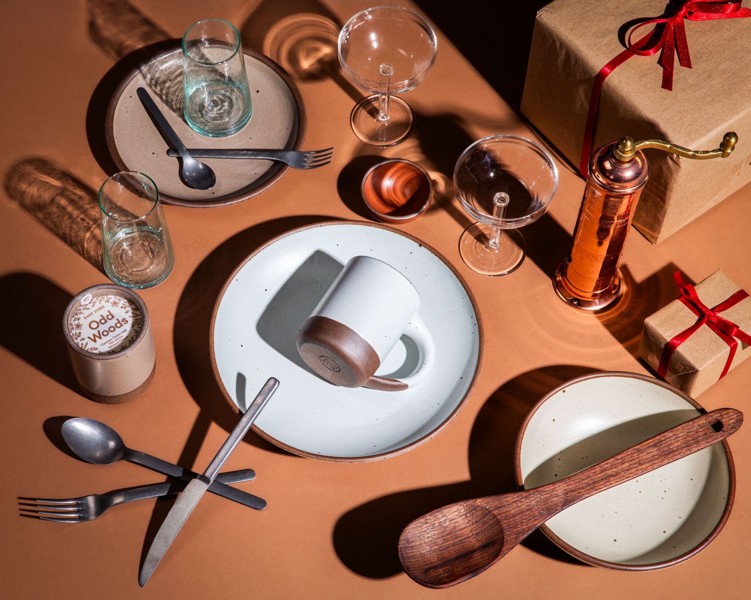 A flatlay of different dinnerware items including ceramic plates, wooden spoon, flatware, martini coupes, and a copper pepper grinder. In the corner is a kraft paper wrapped present with a red ribbon on it.