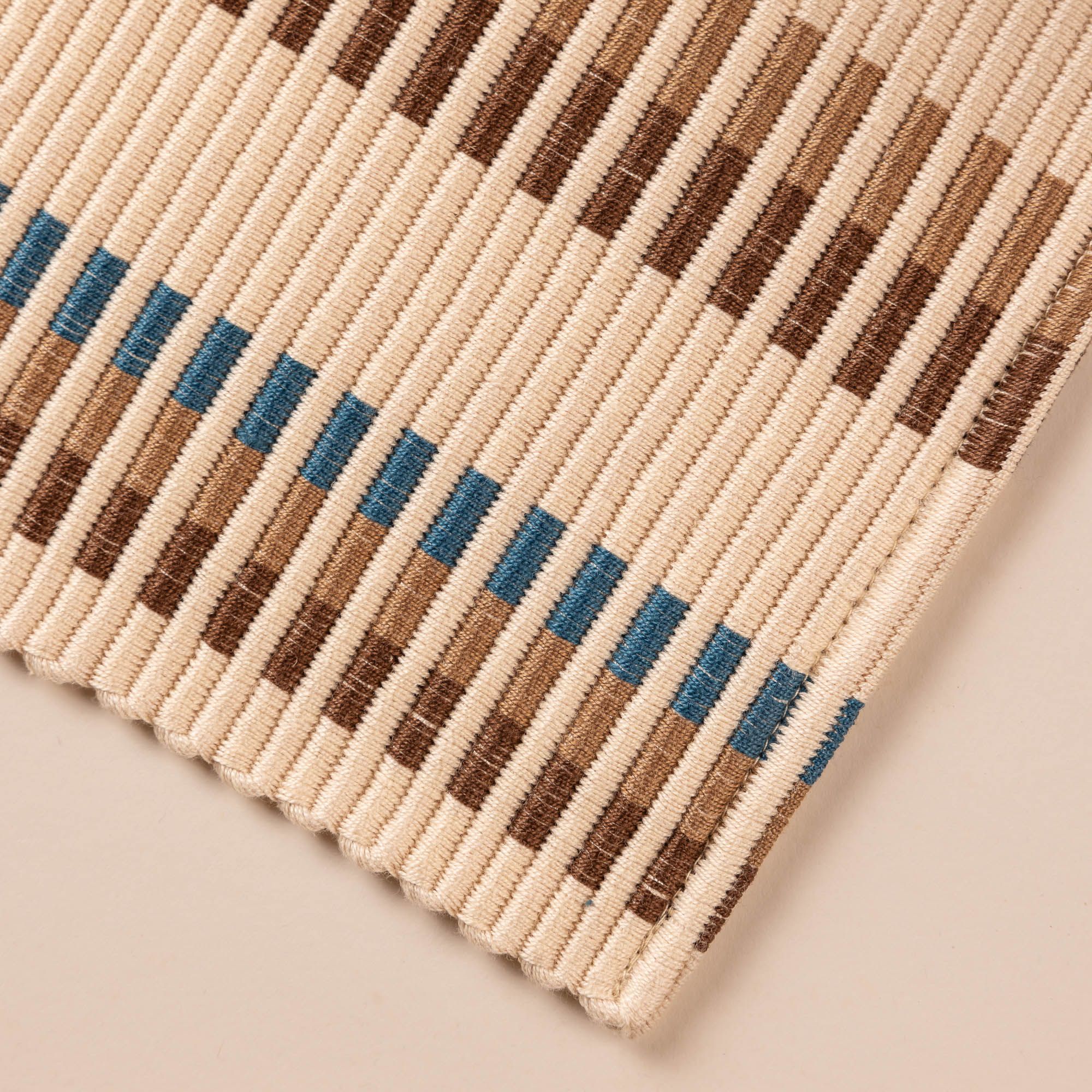 Closeup look at the corner of a rectangle placemat with offset stripe design that is hand woven in cream, brown, tan, and blue colors