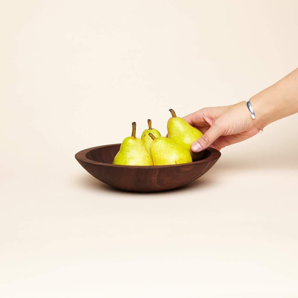 A hand holds one of the pears that sits in a shallow small walnut wood nesting bowl