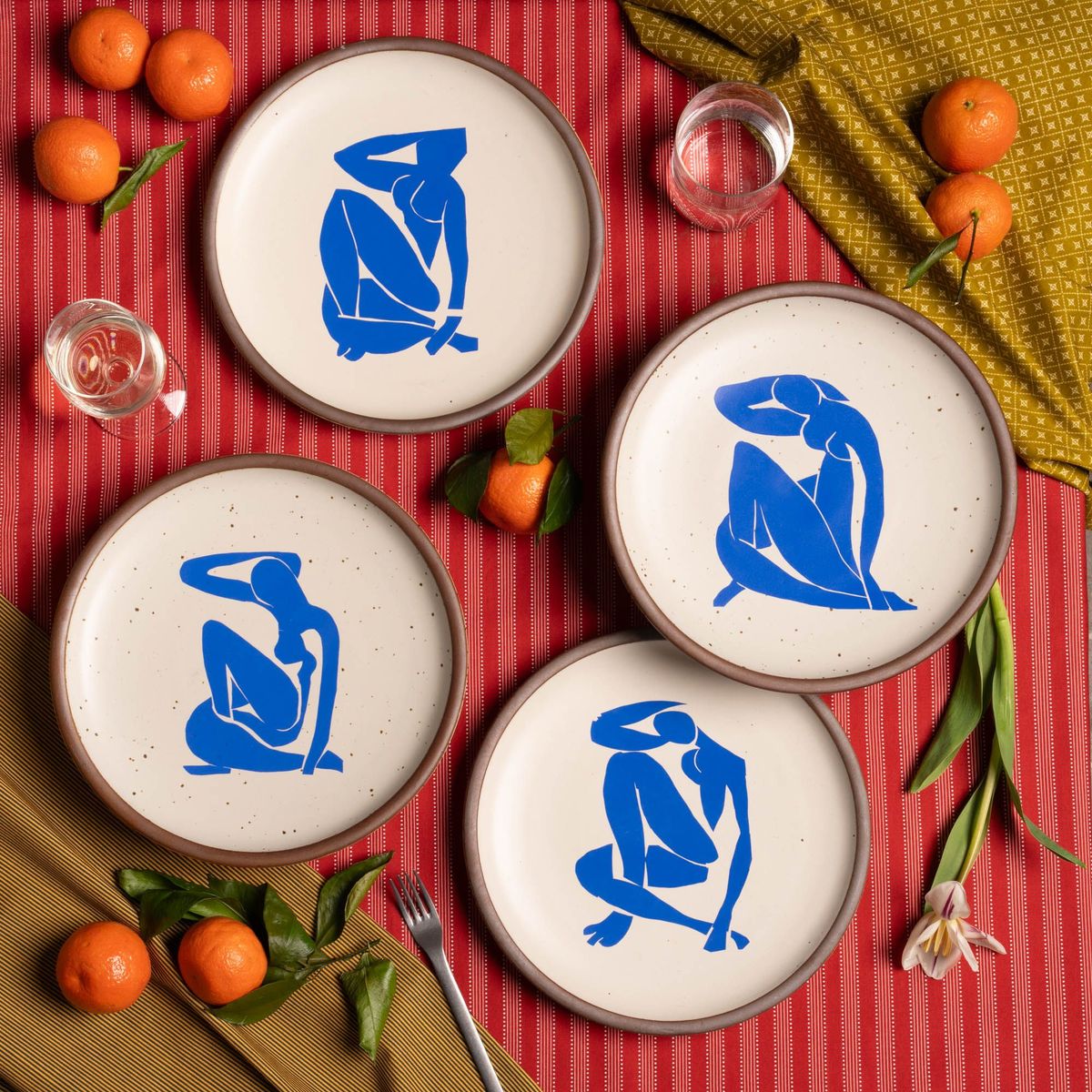 Four ceramic dinner plates in warm off-white colors and unglazed rim, each with a different blue decal from the Nu Bleu series by Henri Matisse. The plates are sitting on a red tablecloth with silverware, flowers, oranges, and glasses.