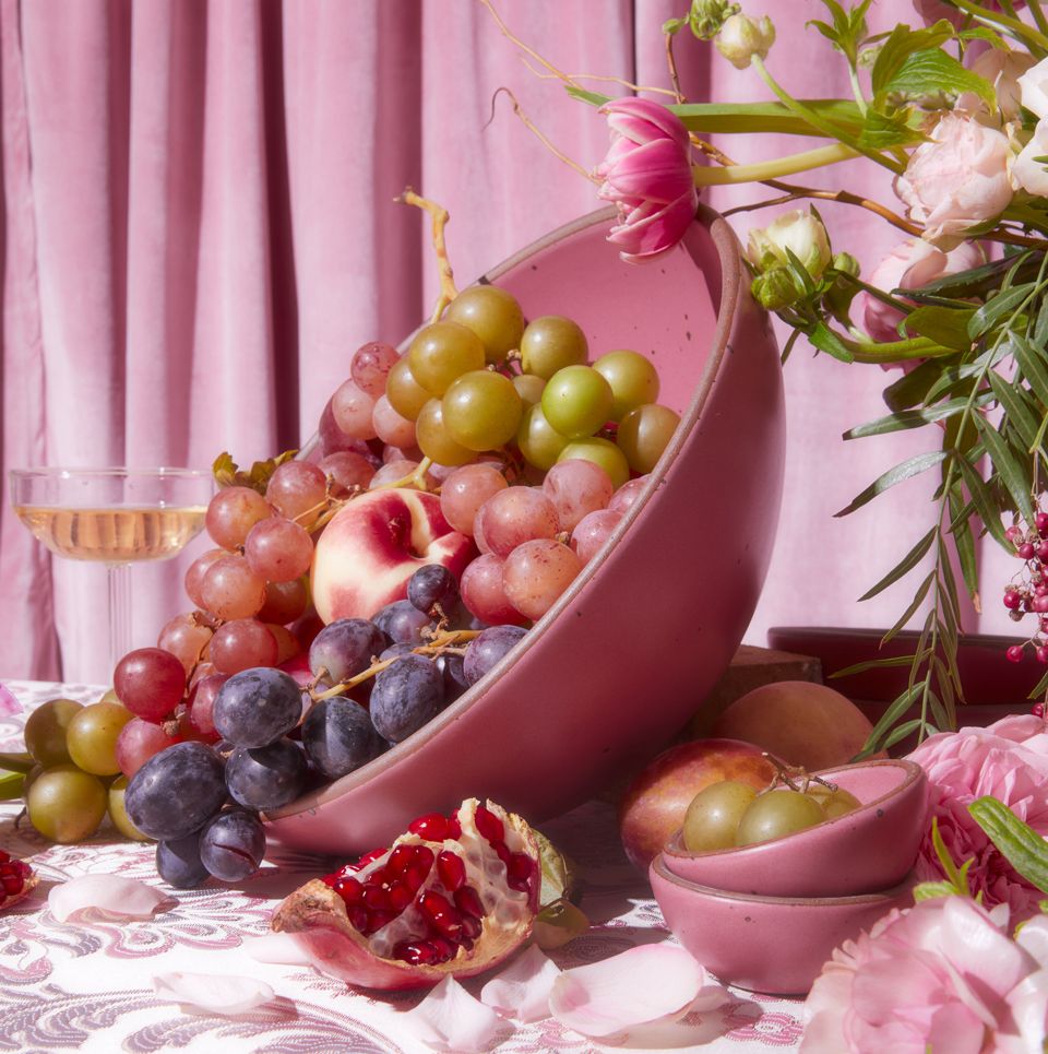 A large pink bowl is tipped on its side and filled with varying colors of grapes and other fruits. A flower arrangement, pink fabric background, and champagne glass is surrounding the bowl.