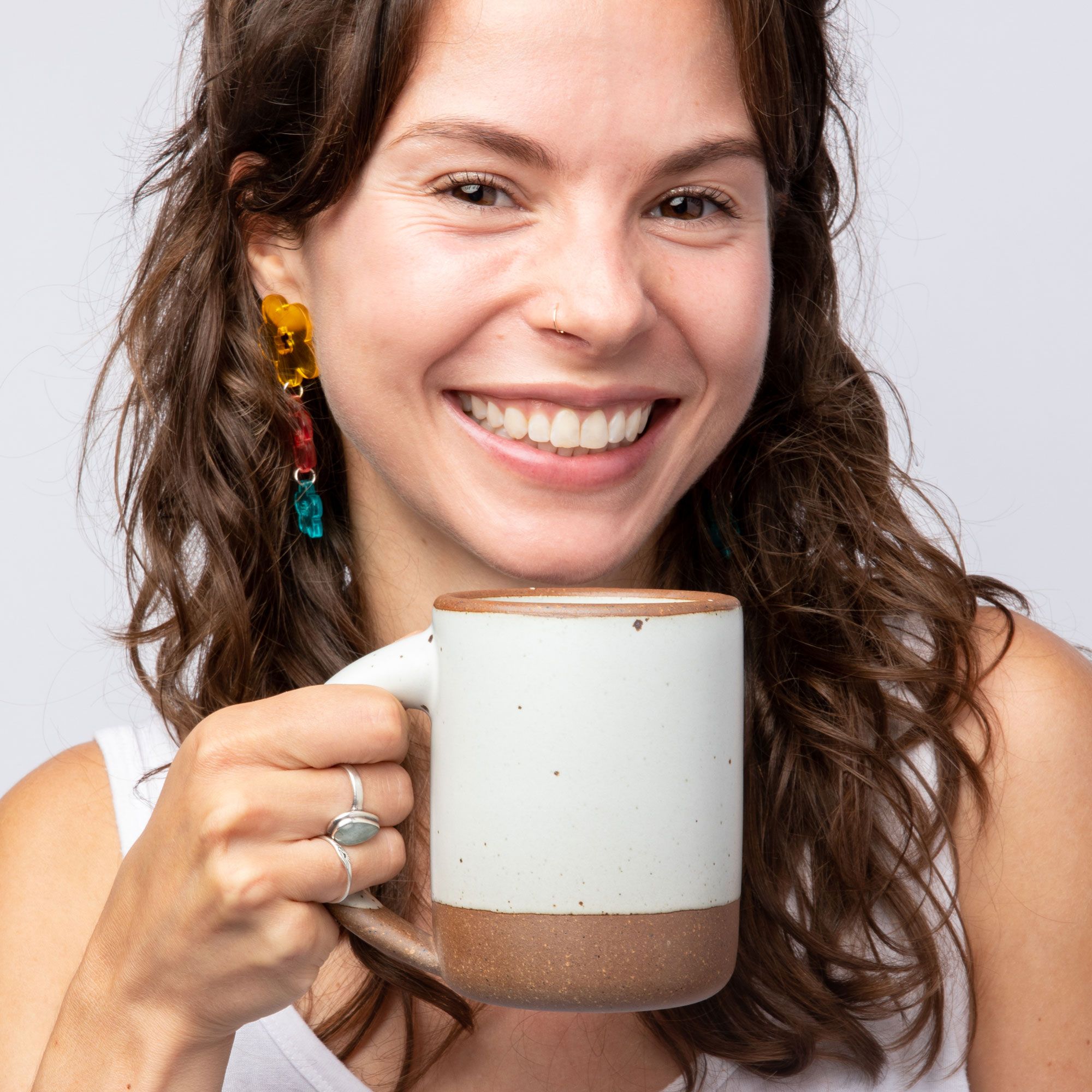 A woman smiling and holding a medium sized ceramic mug with handle in a cool white color featuring iron speckles and unglazed rim and bottom base.