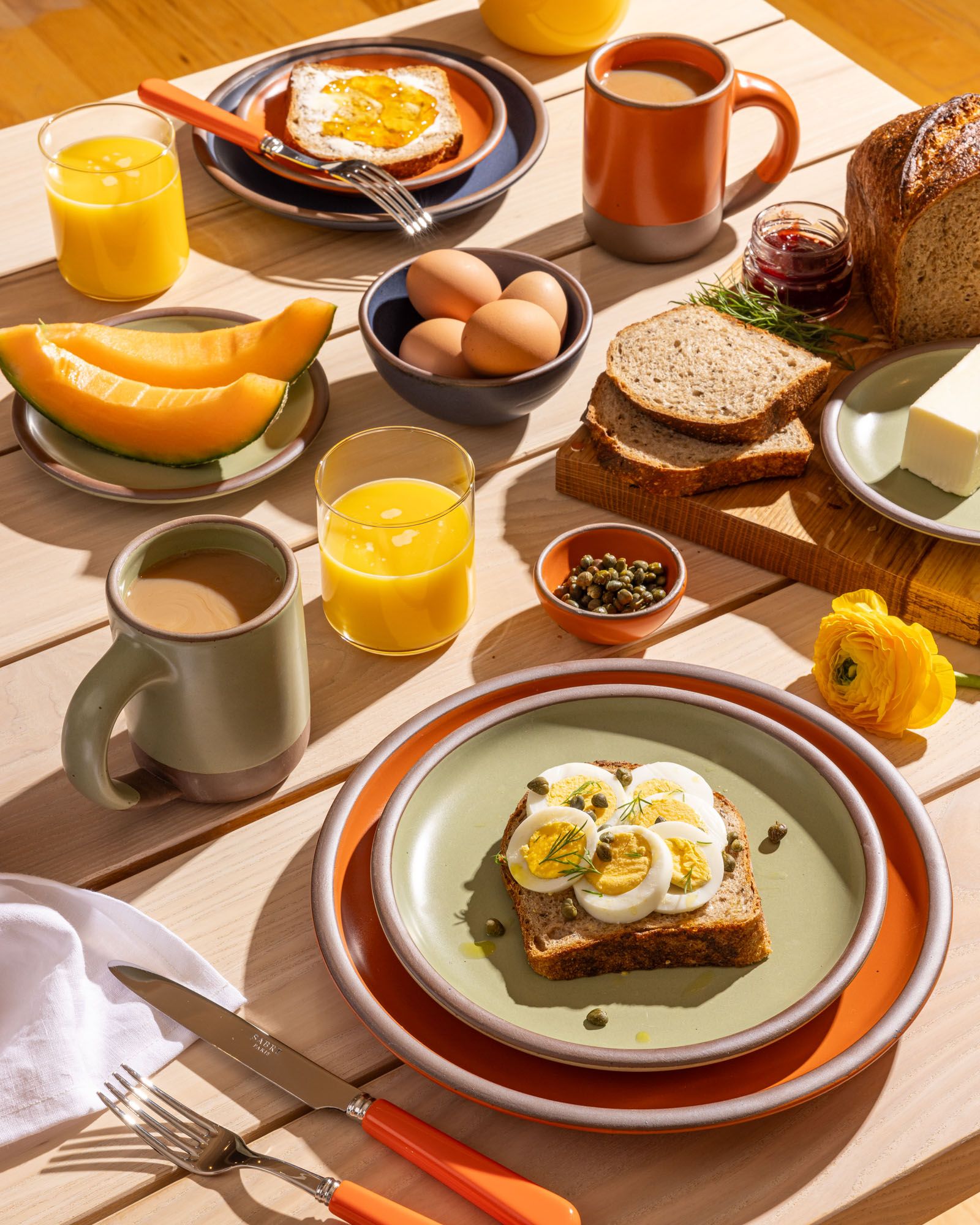 On a wooden table, there is a breakfast spread plated on ceramic pottery in sage green, bold orange, and muted navy colors. Surrounding are juice glasses filled with orange juice, bold orange handled flatware, a wooden cutting board topped with bread, butter, and jam.