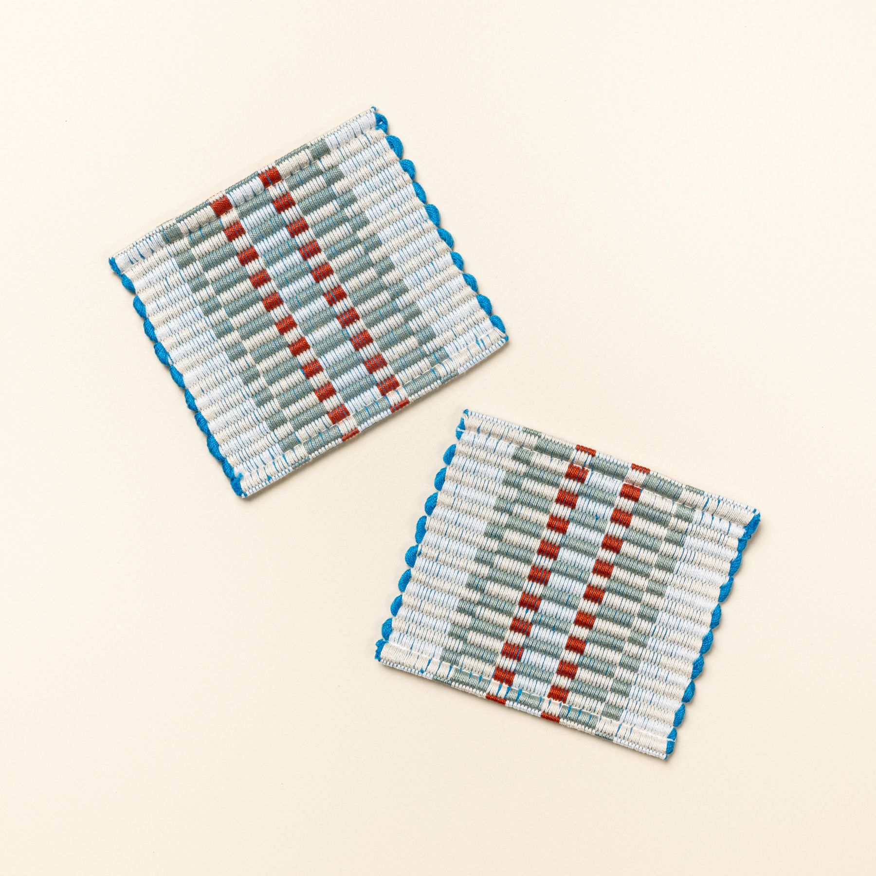 Two square coasters that are hand woven in a striped rectangular grid design in sage, orange, and blue colors