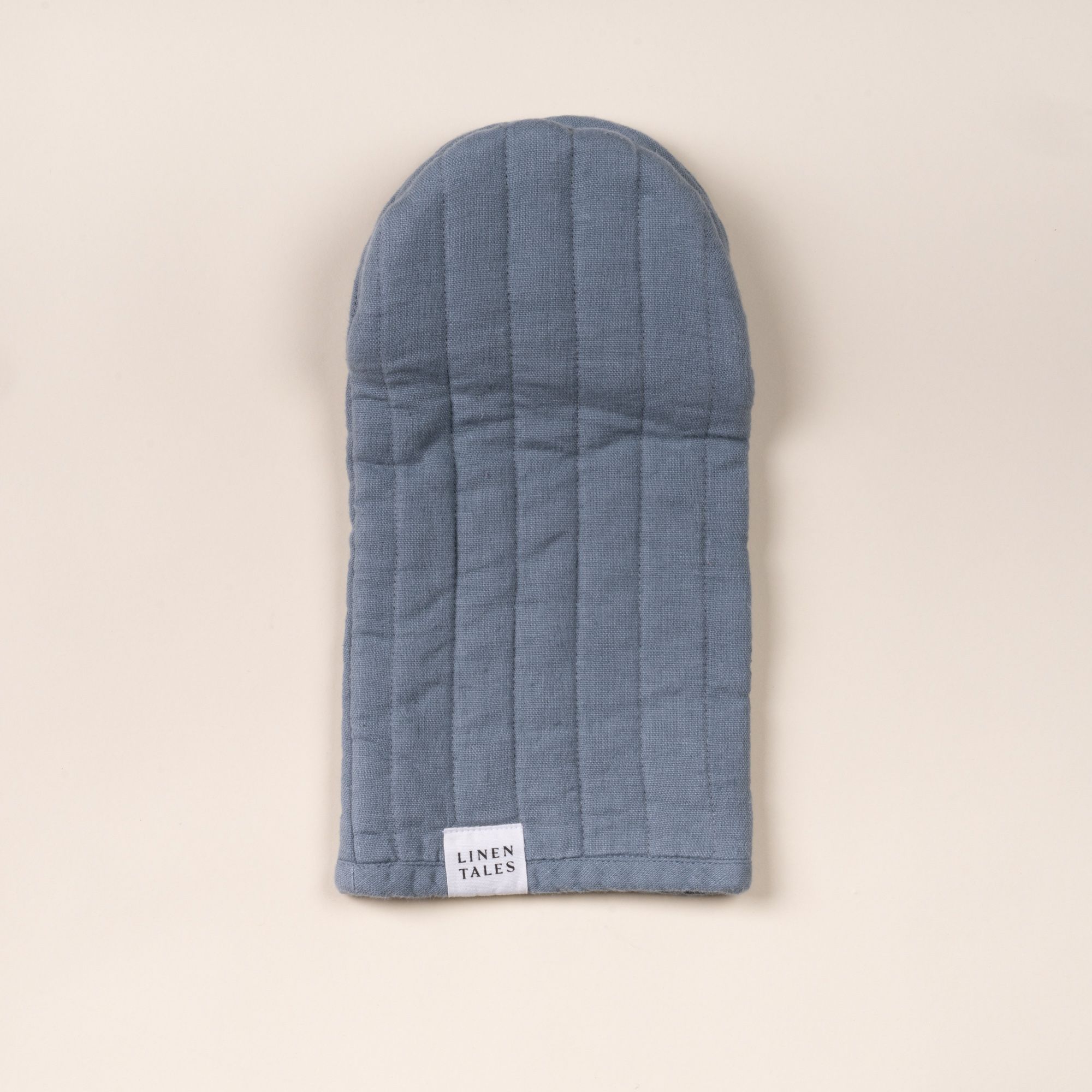 Soft blue linen oven mitt with stitched channels and a small ‘Linen Tales’ label, laid flat on a light background.
