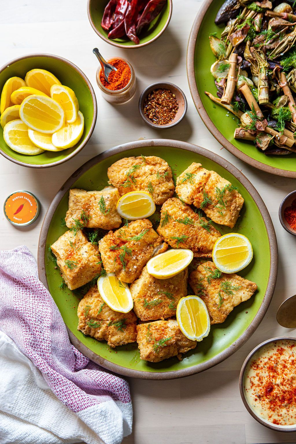 Pan-fried cornmeal cod with lemons on a green dinner plate and surrounded by bowls of ingredients and side dishes.
