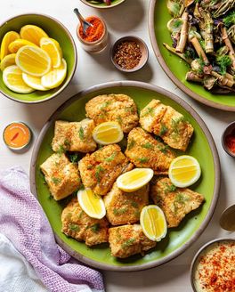 Pan-fried cornmeal cod with lemons on a green dinner plate and surrounded by bowls of ingredients and side dishes.