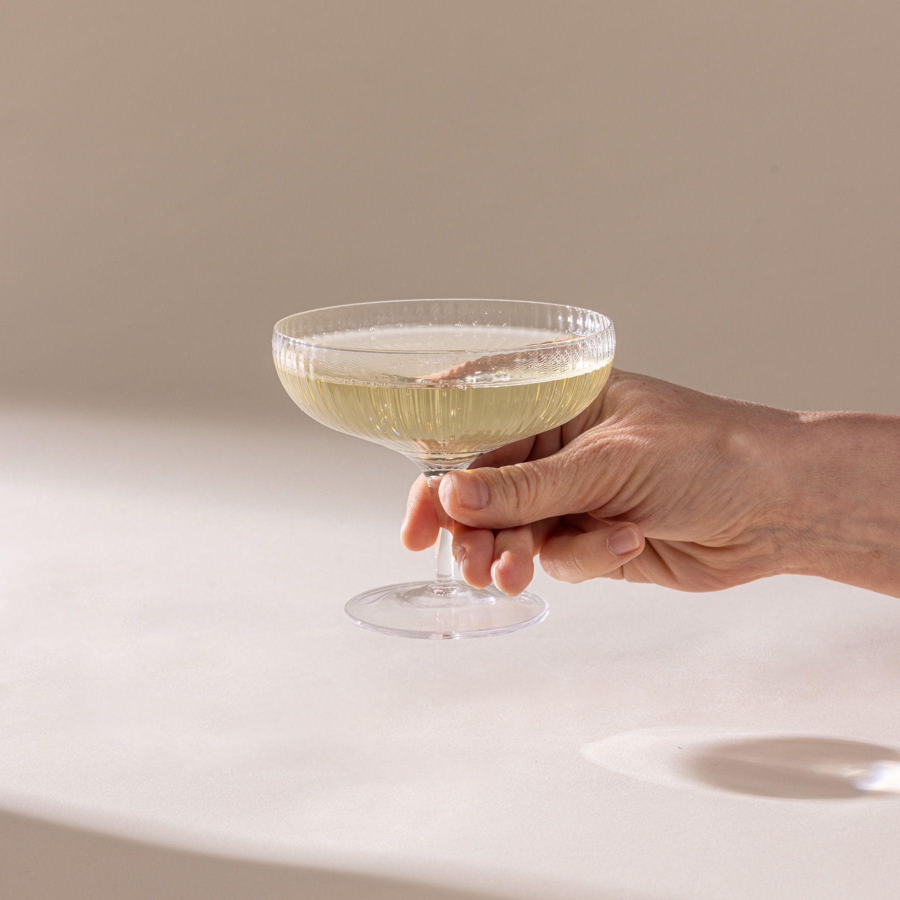 A hand holds a delicate ribbed short coupe glass in the traditional style, filled with champagne