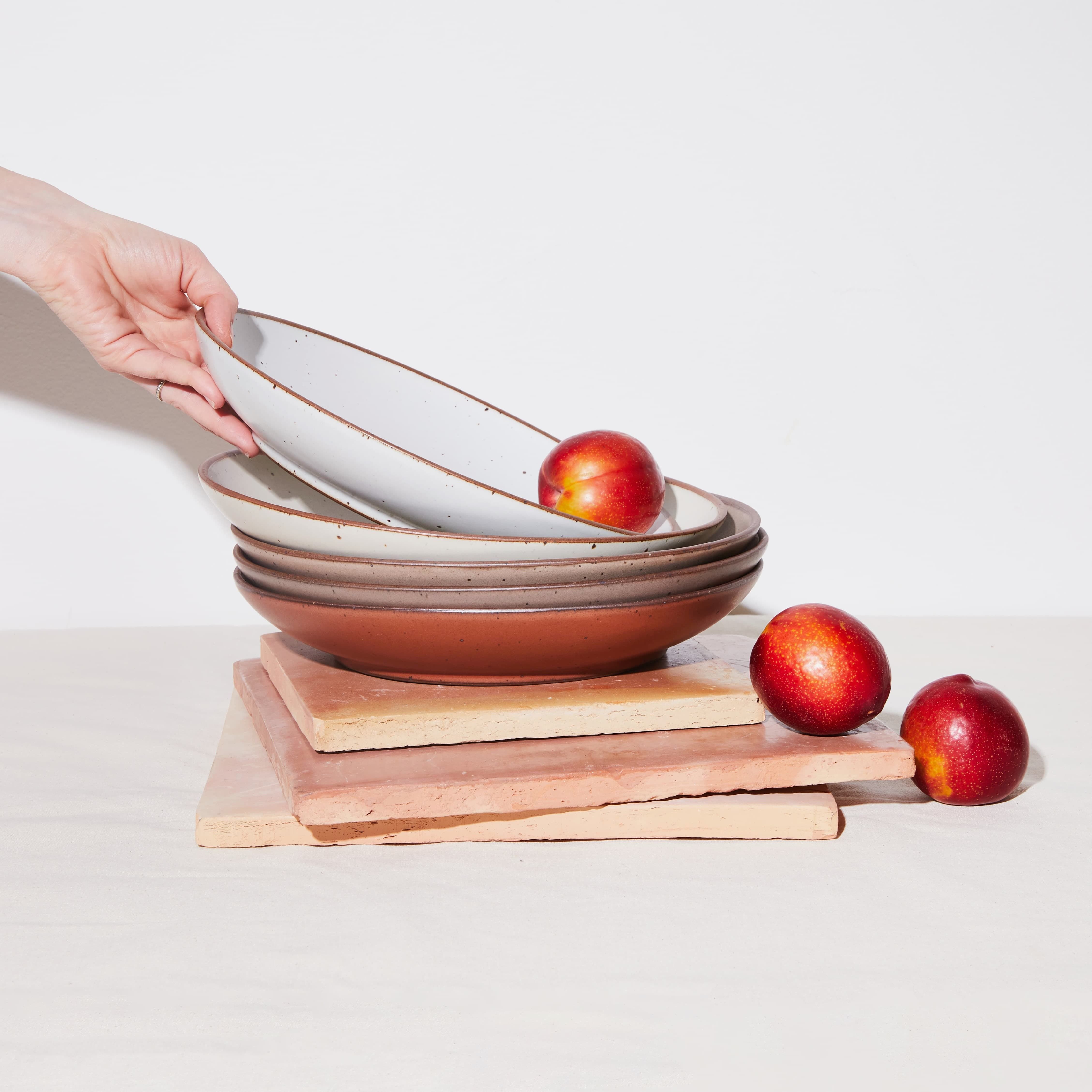 A stack of ceramic coupe plates in terracotta, tan, and white colors with apples falling out of them