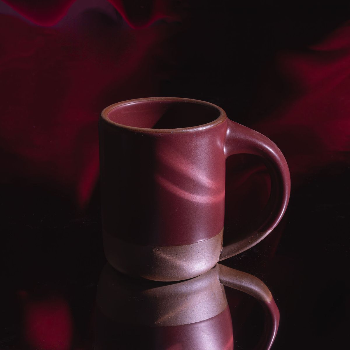 A medium sized ceramic mug with handle in a plum color in a moody setting with reflective light