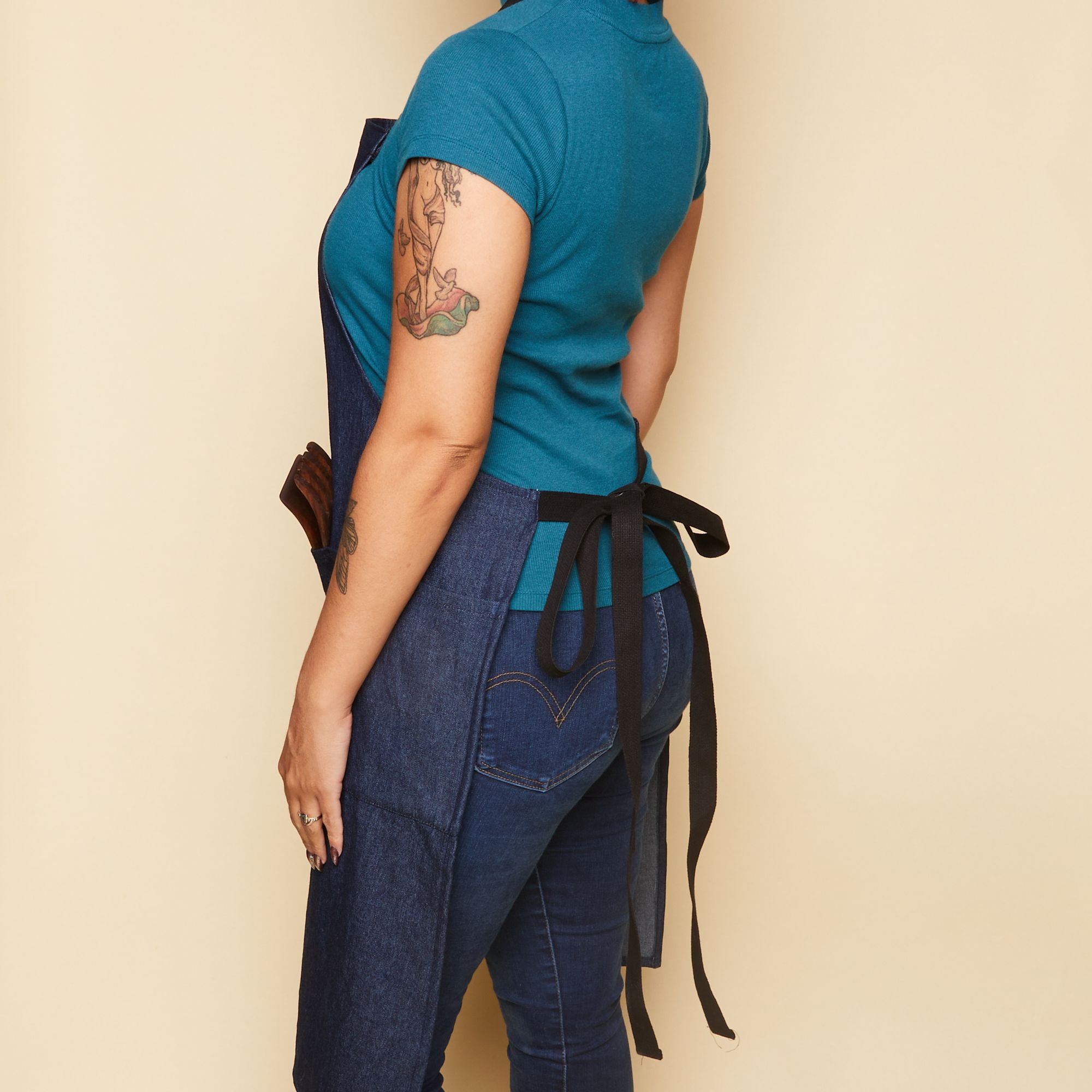 Model smiling wearing a blue shirt and jeans with a long denim blue apron over it that is tied in the back