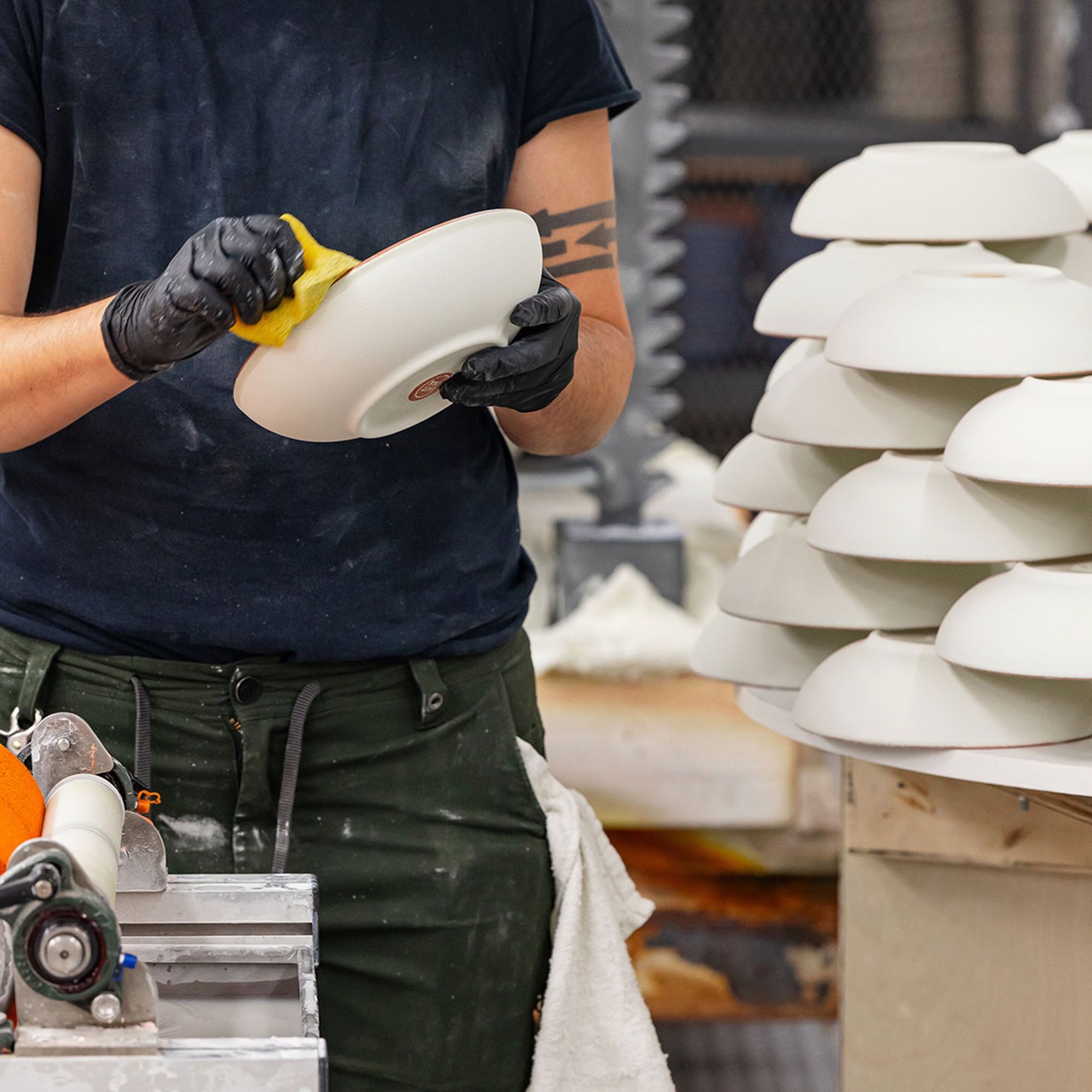 In a factory setting, a person is holding a ceramic bowl in progress and wiping away excess glaze on the rim. A large stack of bowls is next to them.