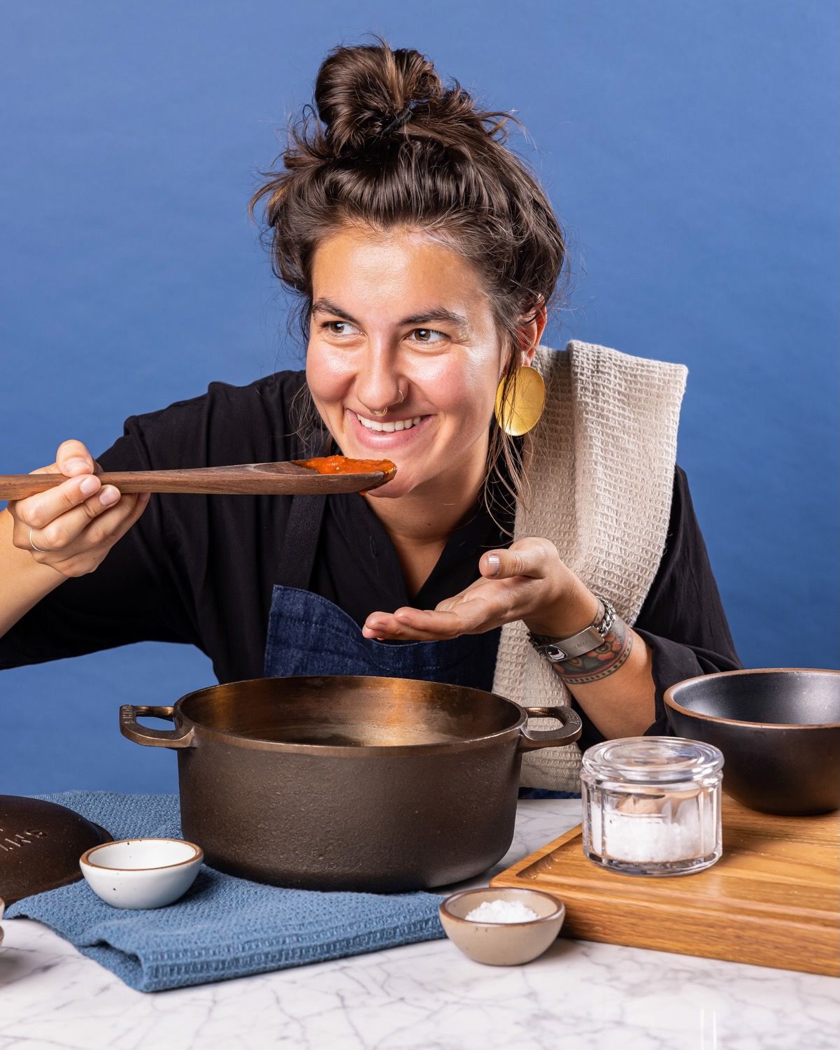 A person is smiling behind a large dutch oven and holding a large wooden spoon with sauce up to their mouth to taste.