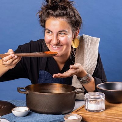 A person is smiling behind a large dutch oven and holding a large wooden spoon with sauce up to their mouth to taste.