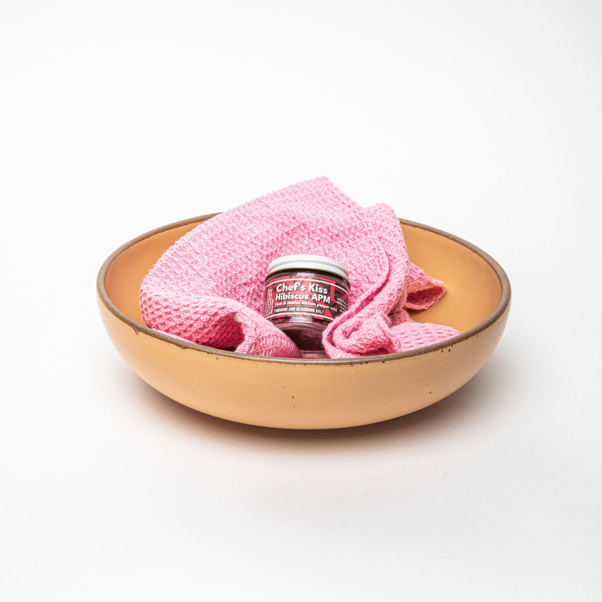 A large serving bowl in a peachy yellow color with a warm pink waffle towel and a small jar with a white top and a label that reads "Chef's Kiss Hibiscus APM" inside