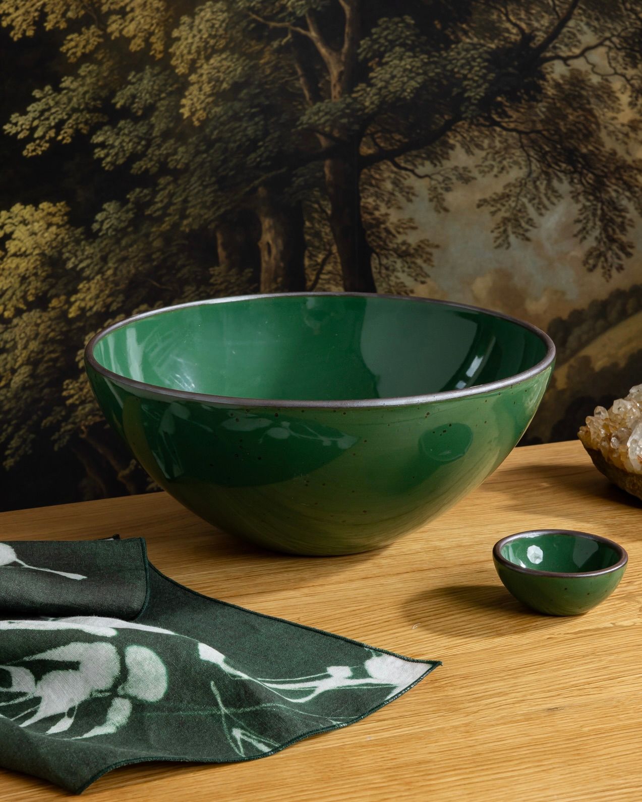 Glossy emerald green ceramic serving bowl with tiny matching bowl on a wooden table, styled with a napkin and crystal accent.