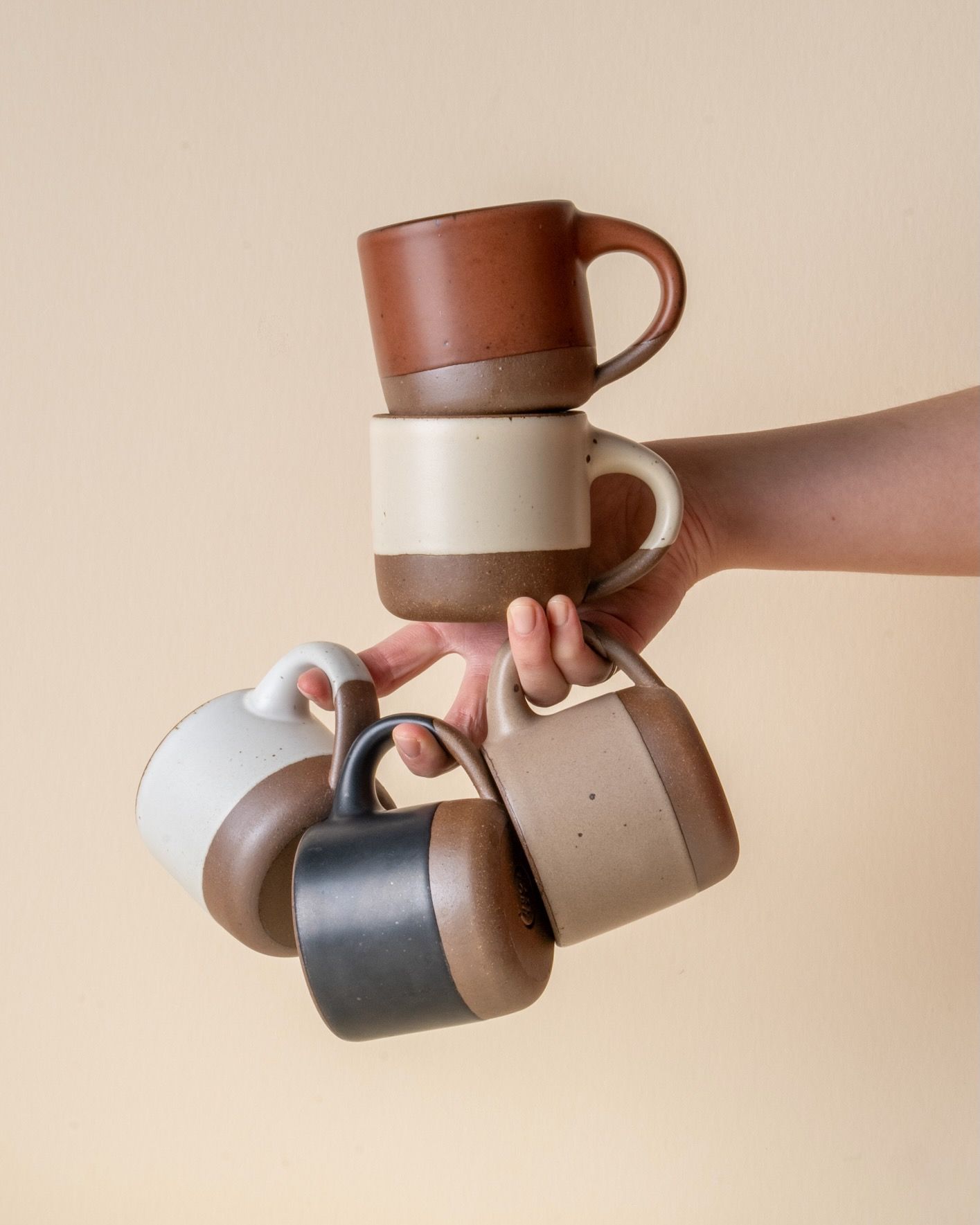 A hand holds out small ceramic mugs on a few fingers and balances 2 small ceramic mugs on the palm of their hand.