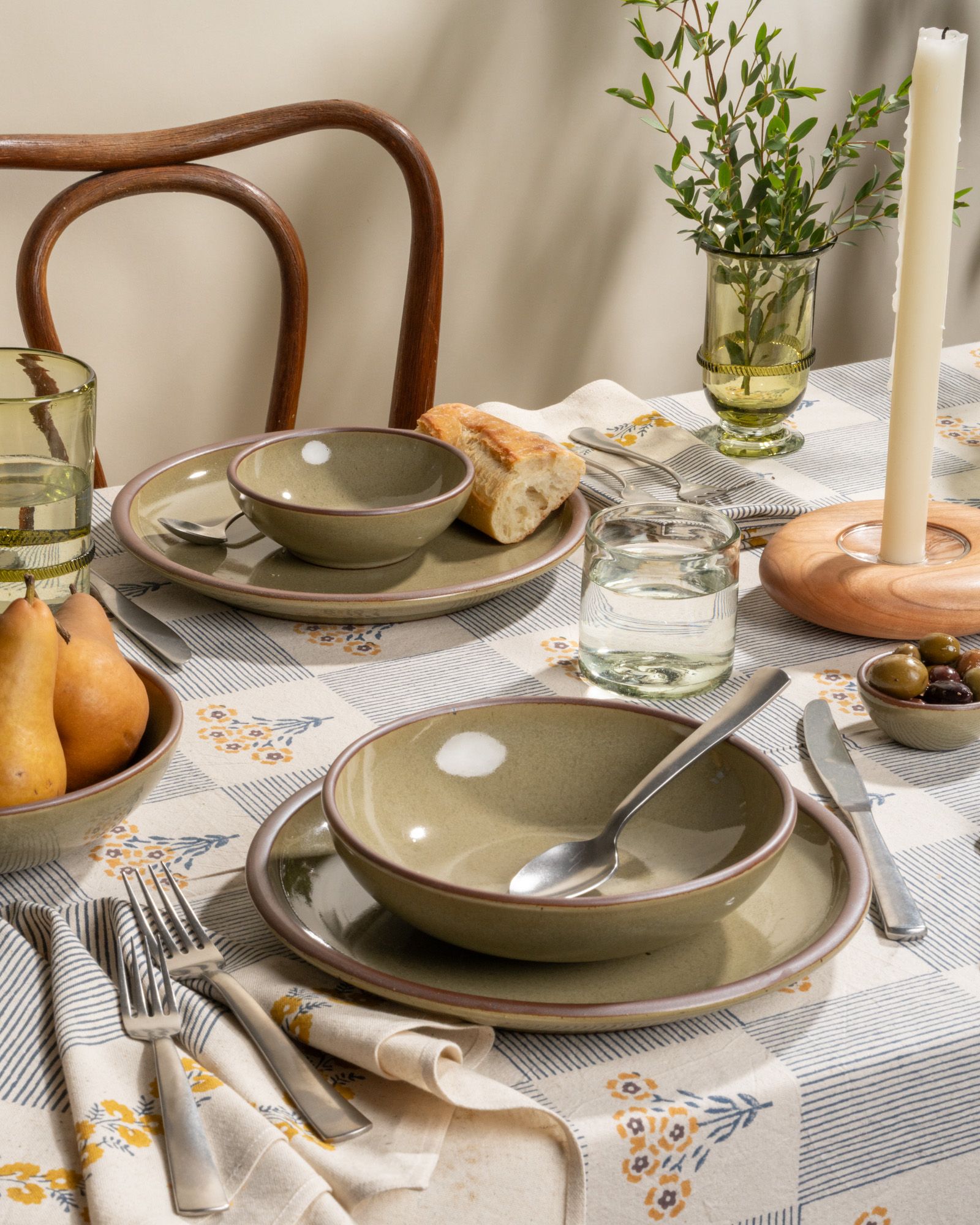 Stoneware grey-green ceramic dinnerware beside taper candles, banded green glass vase, and glassware on a floral tablecloth.