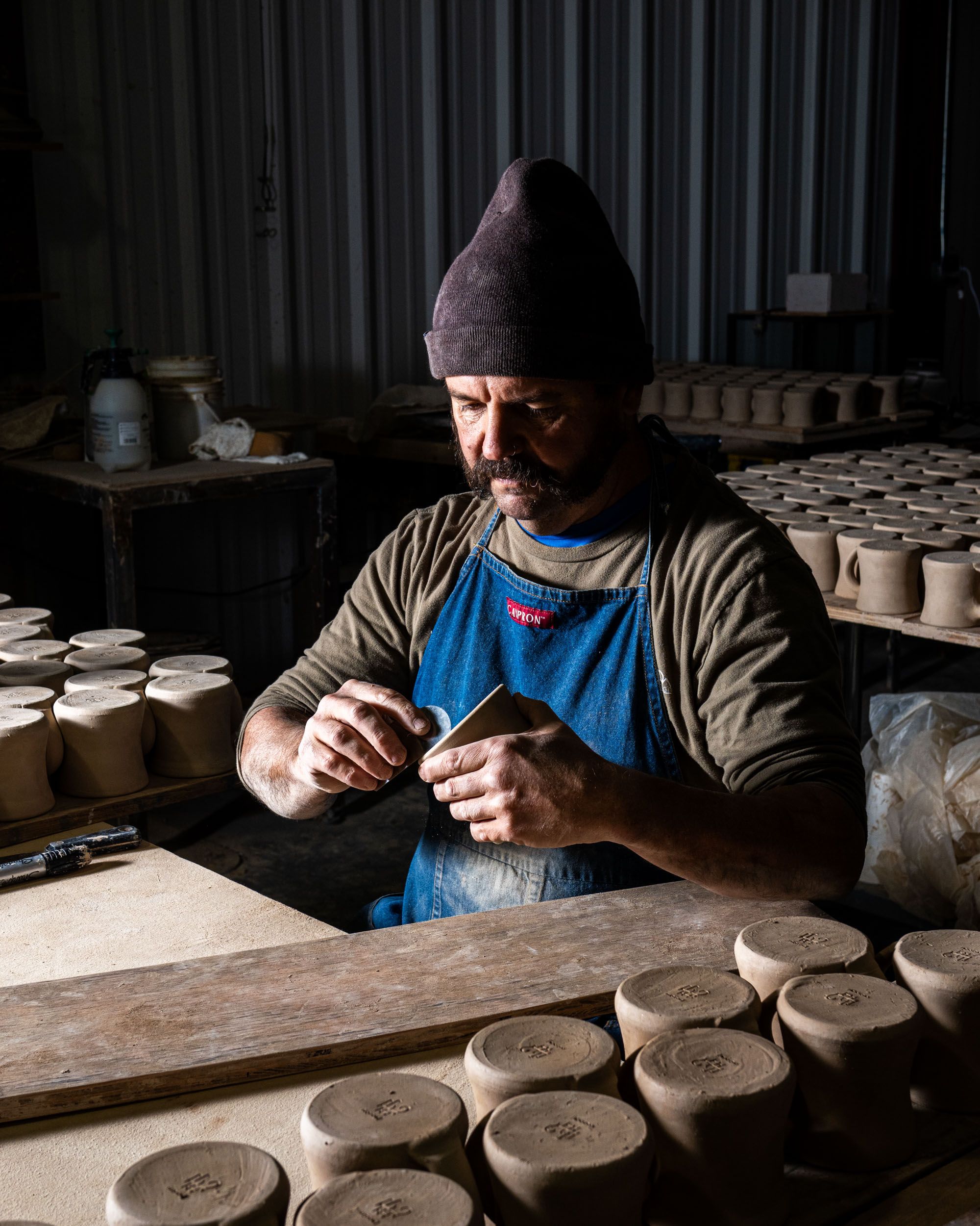 In a pottery studio setting, a man is forming the bottom base of a Diner Mug. Surrounding him are hundreds of mugs.