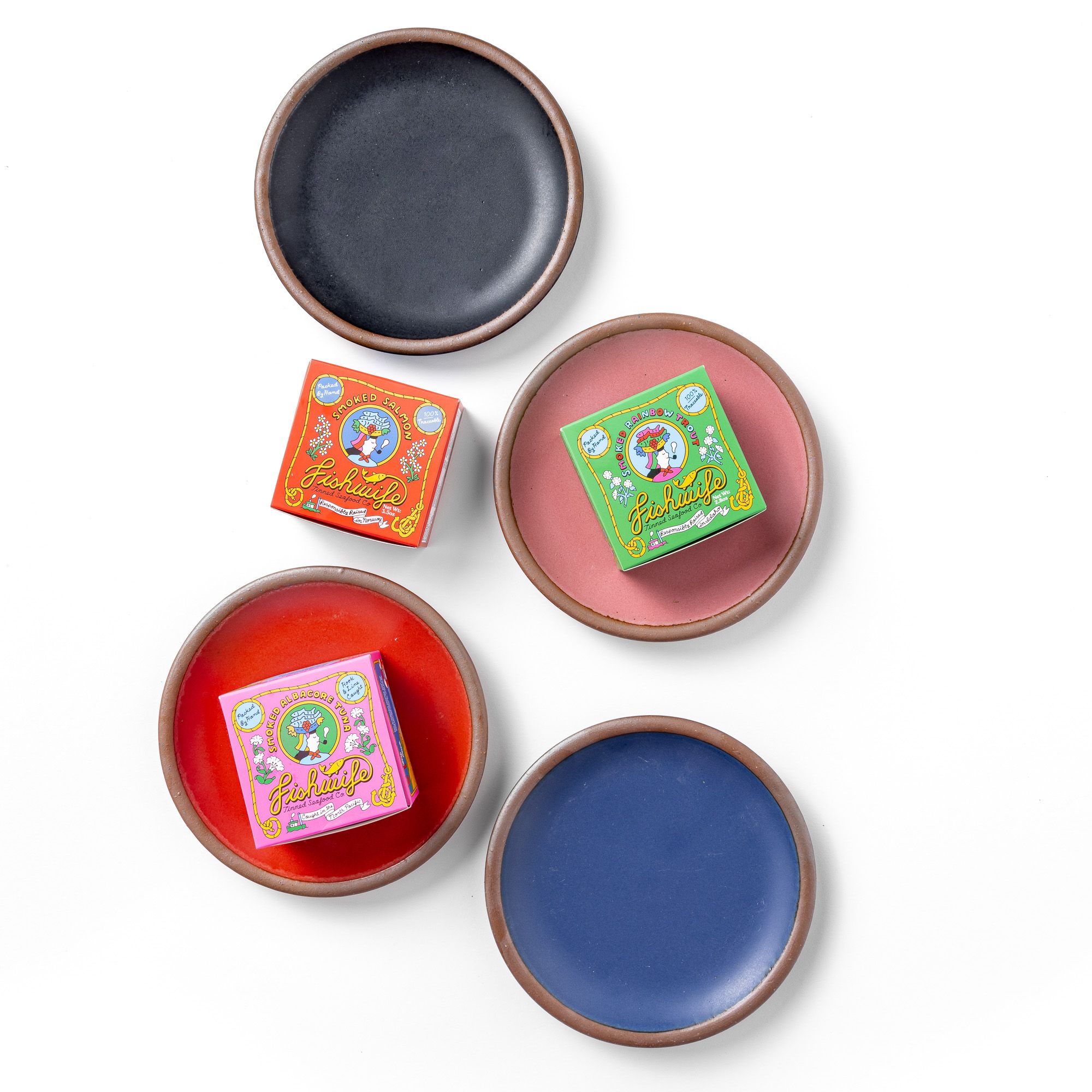 4 small ceramic plates are next to each other in blue, red, pink and black colors. On top and beside them are 3 boxes of tinned fish in pink, orange, and green colors